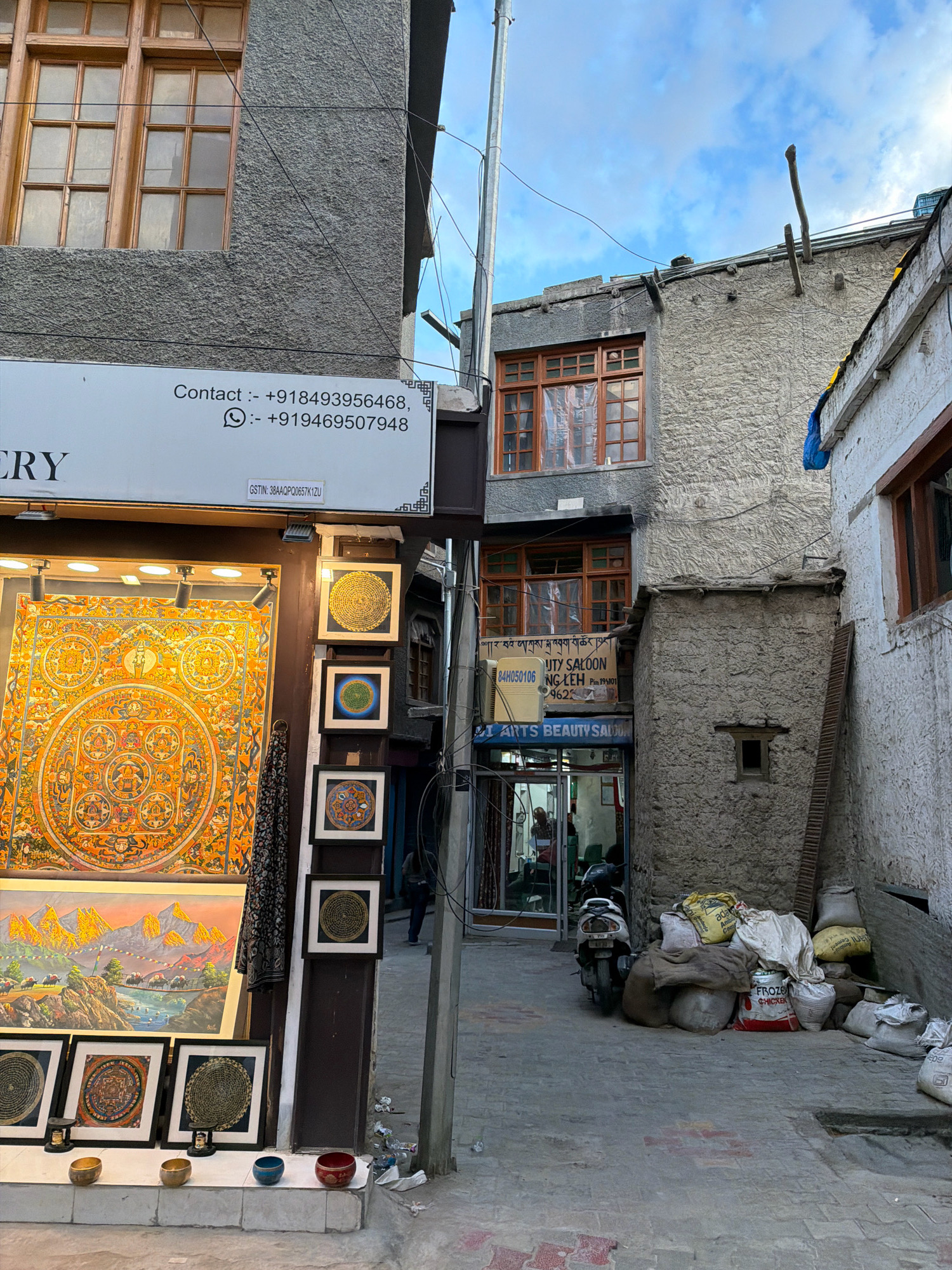 The image depicts a narrow street in Leh, India, likely a commercial area. On the left, a shop window displays colorful artwork, including a large mandala and framed paintings of mountains and Tibetan Buddhist art. Above the shop, a sign provides contact information including phone numbers and a GSTIN. The building on the left has a grey textured facade and a wooden-framed window.
To the right, a two-story building with a rough, grey plastered exterior is visible. This building has wooden-framed windows with some windows covered by plastic sheeting. Below the windows, a sign for "ARTS BEAUTY SALOON" is displayed. A narrow alleyway leads further into the scene, with a scooter parked near the entrance to the salon. Bags of goods, some appearing to be cement or rice sacks, are piled against the wall on the right side of the alley. The ground is paved with interlocking bricks. The sky above is partly cloudy. A person can be seen walking in the background of the alleyway.