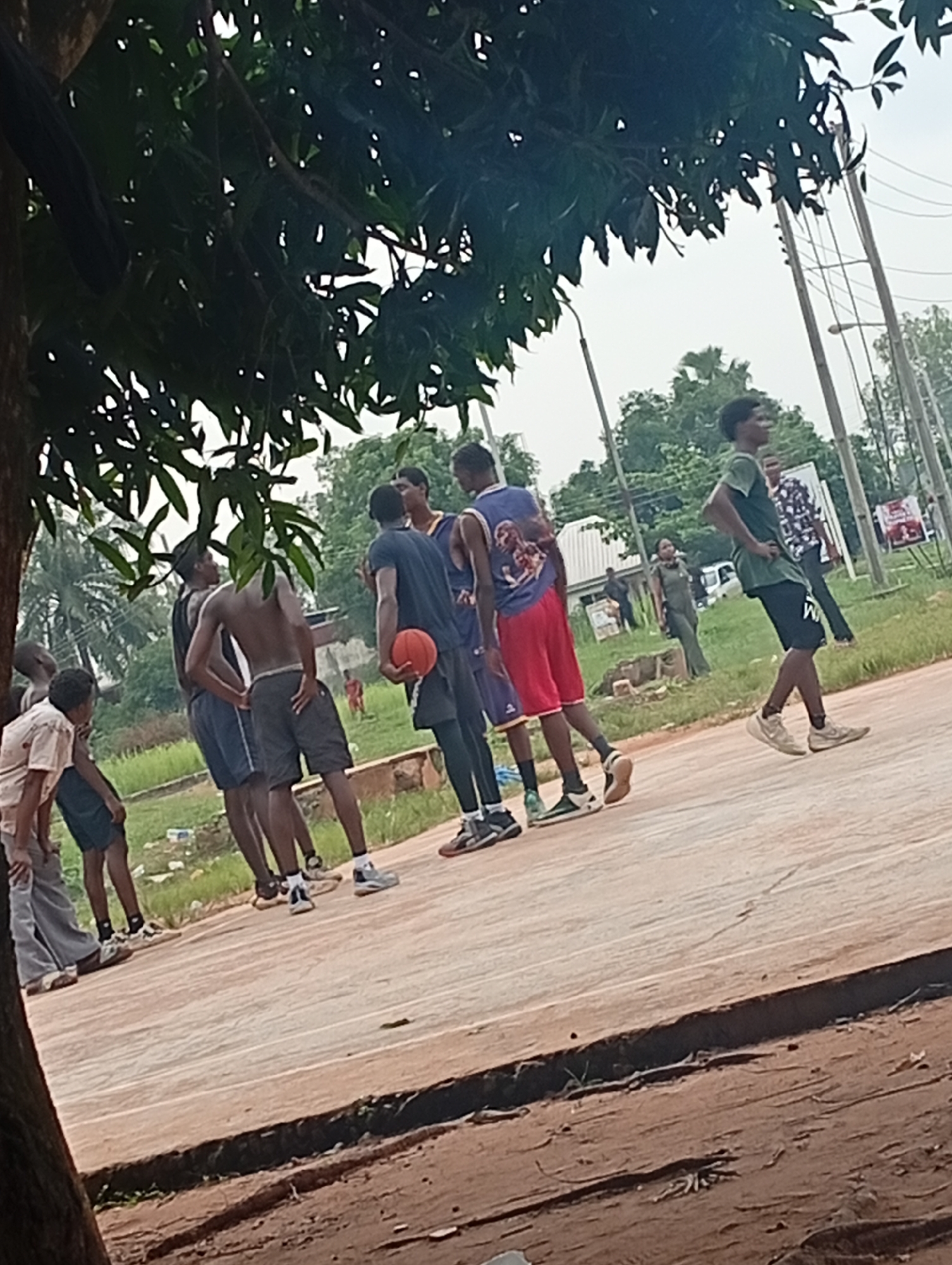 In Ukukwa, Nigeria, a group of young men are gathered on an outdoor basketball court, seemingly engaged in a game or practice. The court surface is a mix of concrete and dirt. The individuals are dressed in casual athletic wear, with some shirtless and others in jerseys and shorts. One person is holding a basketball, indicating an ongoing activity. The background reveals a lush green environment with trees and vegetation, characteristic of the Nigerian landscape. Utility poles and wires are visible in the distance, suggesting the presence of infrastructure within the community. The lighting suggests it is daytime, likely overcast or with diffused sunlight, as indicated by the soft shadows.