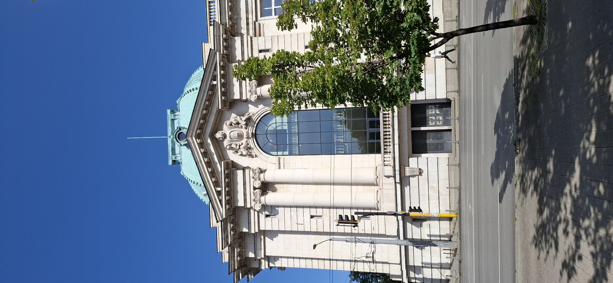 The image captures a frontal view of the National Theatre "Ivan Vazov" in Sofia, Bulgaria, under a clear blue sky. The building features an ornate facade with classical architectural elements, including Corinthian columns and sculptures above a large, arched window. A distinctive green dome crowns the structure, topped with a slender spire. In the foreground, a tree with vibrant green leaves partially obscures the right side of the building. A street with a pedestrian walkway and traffic lights runs in front of the theatre. A single pedestrian, dressed in a white t-shirt and dark pants, walks along the sidewalk to the right of the frame. The sidewalk is paved with dark gray tiles and is partially shaded by the tree's canopy. A traffic light and utility pole stand to the left of the theatre.