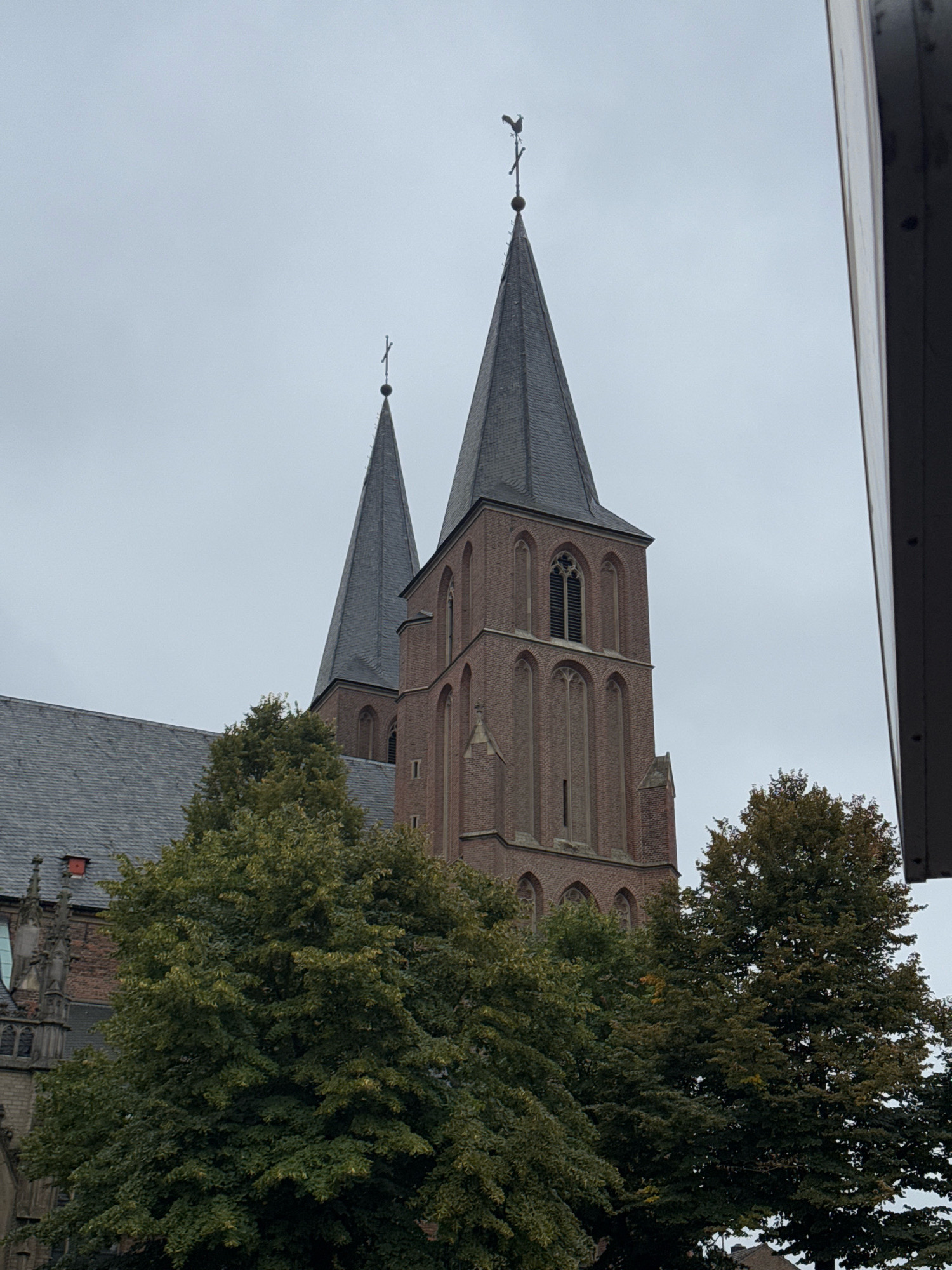 The image shows the towers of the Church of the Holy Spirit (Heilig Geist Kirche) in Cleves, Germany, under an overcast sky. The church features two tall, pointed spires, one of which is topped with a weather vane shaped like a rooster. The spires are constructed from brick and slate. In the foreground, lush green trees with dense foliage partially obscure the lower portions of the church. The overall scene suggests an exterior view of a historical religious building situated within a town or city.