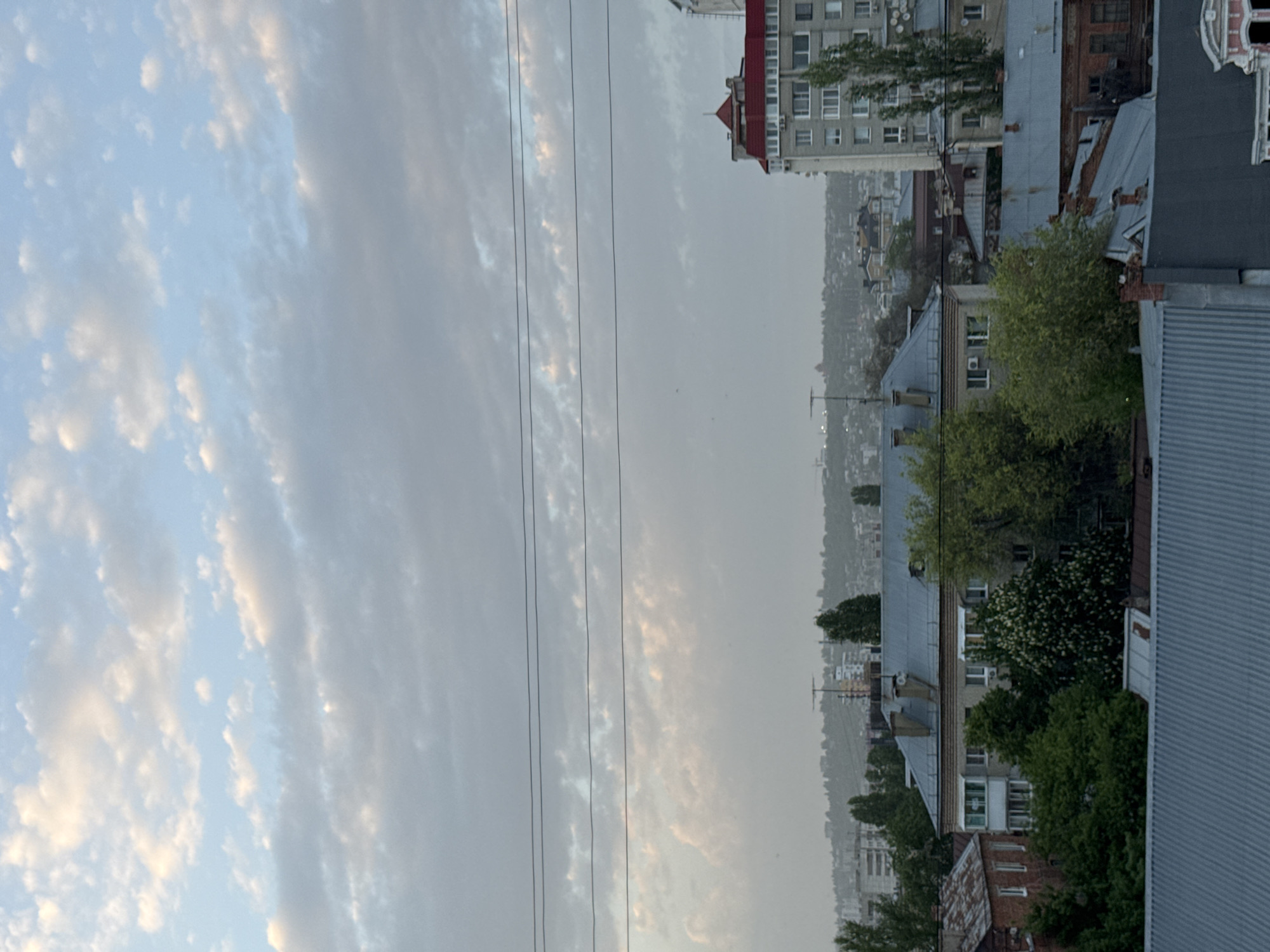 The image depicts a cityscape in Saratov, Russia. The perspective is from an elevated position, looking towards a distant horizon with a dense urban environment in between. The foreground shows rooftops of various buildings, interspersed with trees. In the distance, buildings of different heights and architectural styles are visible, creating a layered effect. The sky is filled with clouds, ranging from light and wispy to more dense and grey. Power lines are seen stretching across the sky in the middle ground.