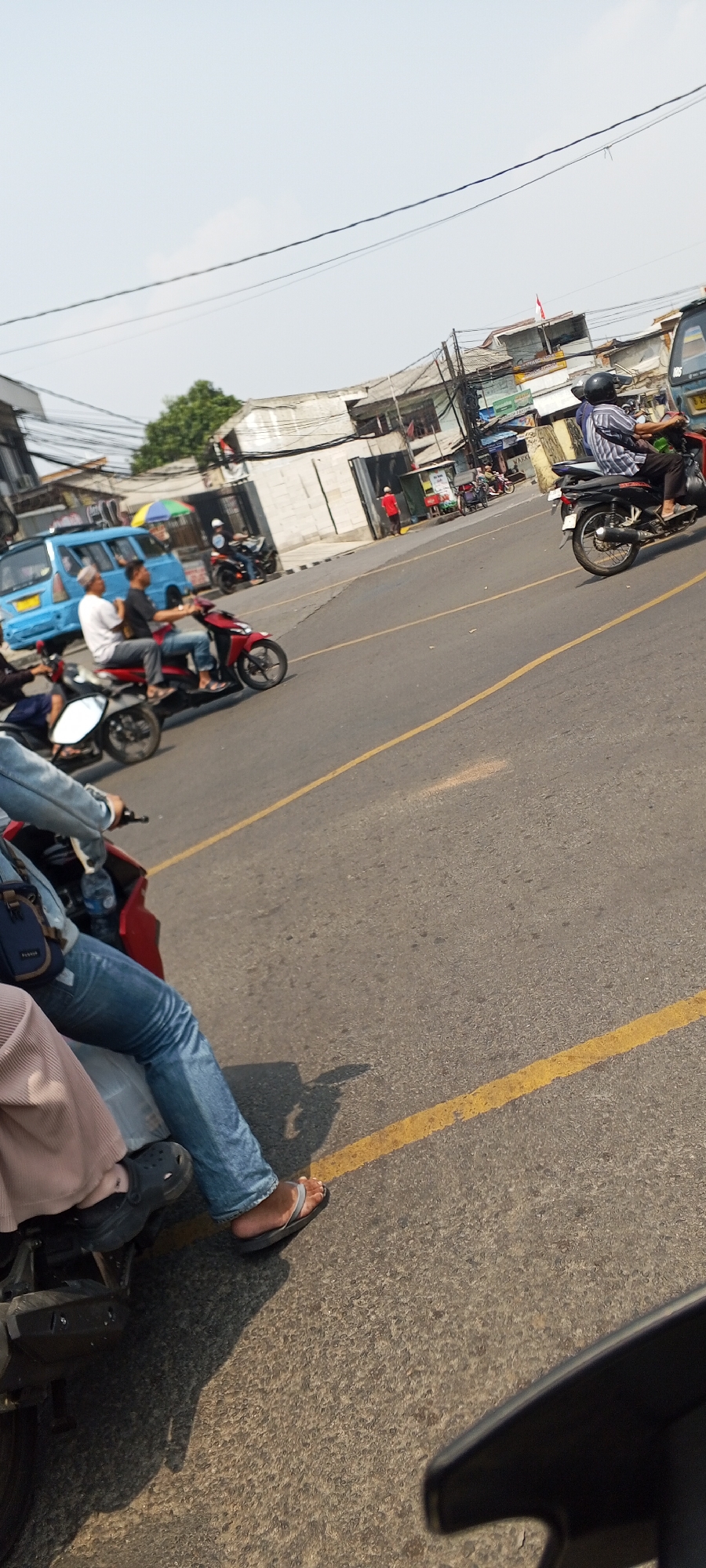 In Depok, Indonesia, on a sunny day, a street scene unfolds with several motorcycles and a blue van. A person wearing a light blue denim jacket and blue jeans rides a red motorcycle, with their left leg extended forward. They wear gray flip-flops. To their right, another motorcycle is partially visible. In the background, a blue van is parked. Further back, a person in a black and white striped shirt and black pants rides a black motorcycle. Other motorcycles and pedestrians are present in the distance. Buildings with varying architectural styles line the street, some appearing weathered. Power lines are visible crisscrossing overhead. The road is asphalt with yellow lane markings. Shadows from the vehicles and people are cast on the road.