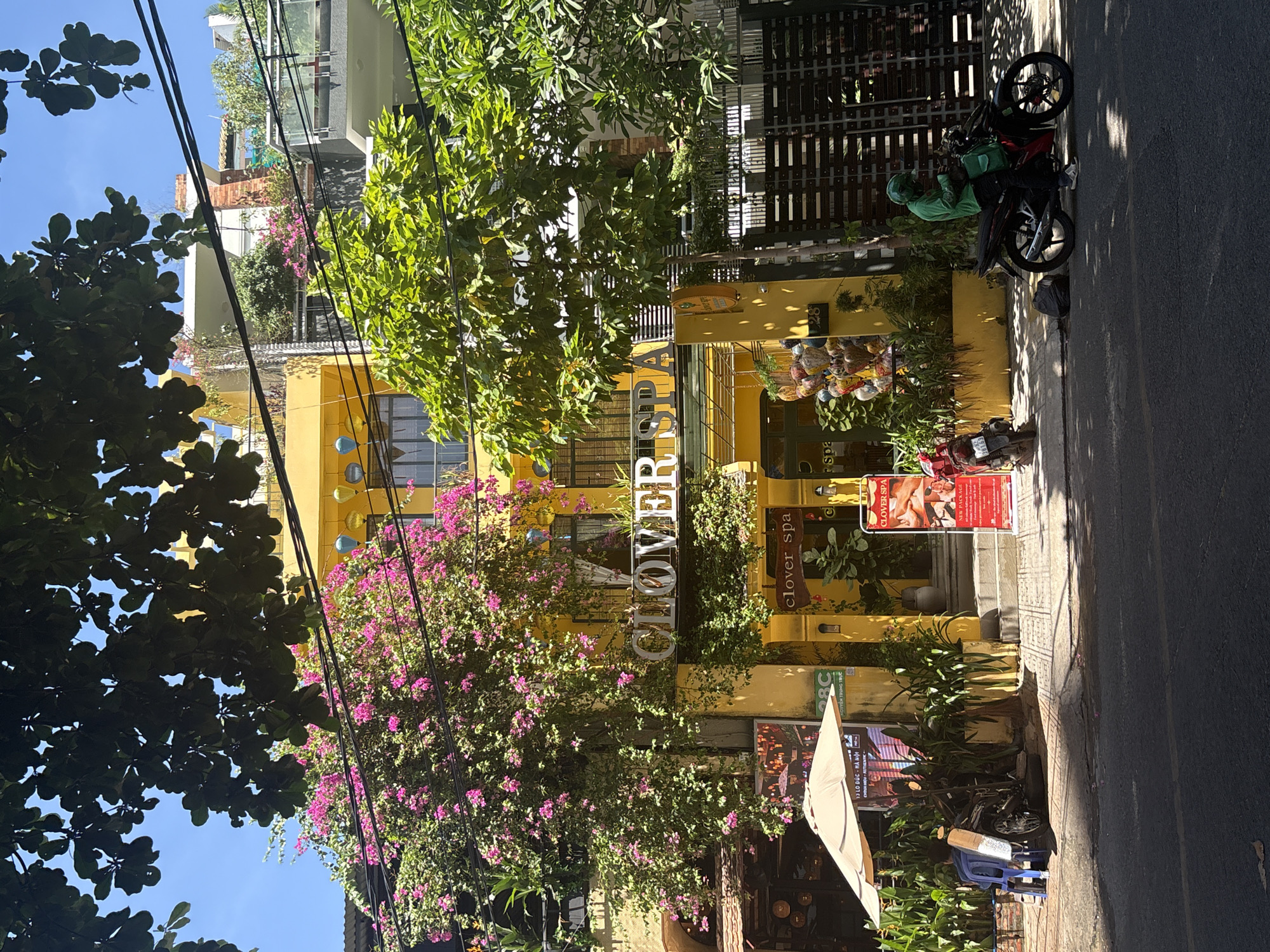 In Nha Trang, Vietnam, a vibrant yellow building with "CLOVER SPA" signage stands prominently, adorned with lush pink bougainvillea. The street in front of the spa is bustling with activity. A person in a green outfit, wearing a helmet, is seated on a black motorcycle, seemingly checking their phone. Nearby, another motorcycle is parked. A person is seated on a blue chair under a large umbrella, facing a stall with various items. Plants line the walkway and stairs leading up to the spa entrance. The building is a multi-story structure with balconies and windows, suggesting it may also house residences or other businesses. The scene is bathed in bright sunlight, casting shadows across the pavement, indicating a clear day. The surrounding environment appears to be an urban street with other buildings visible in the background.