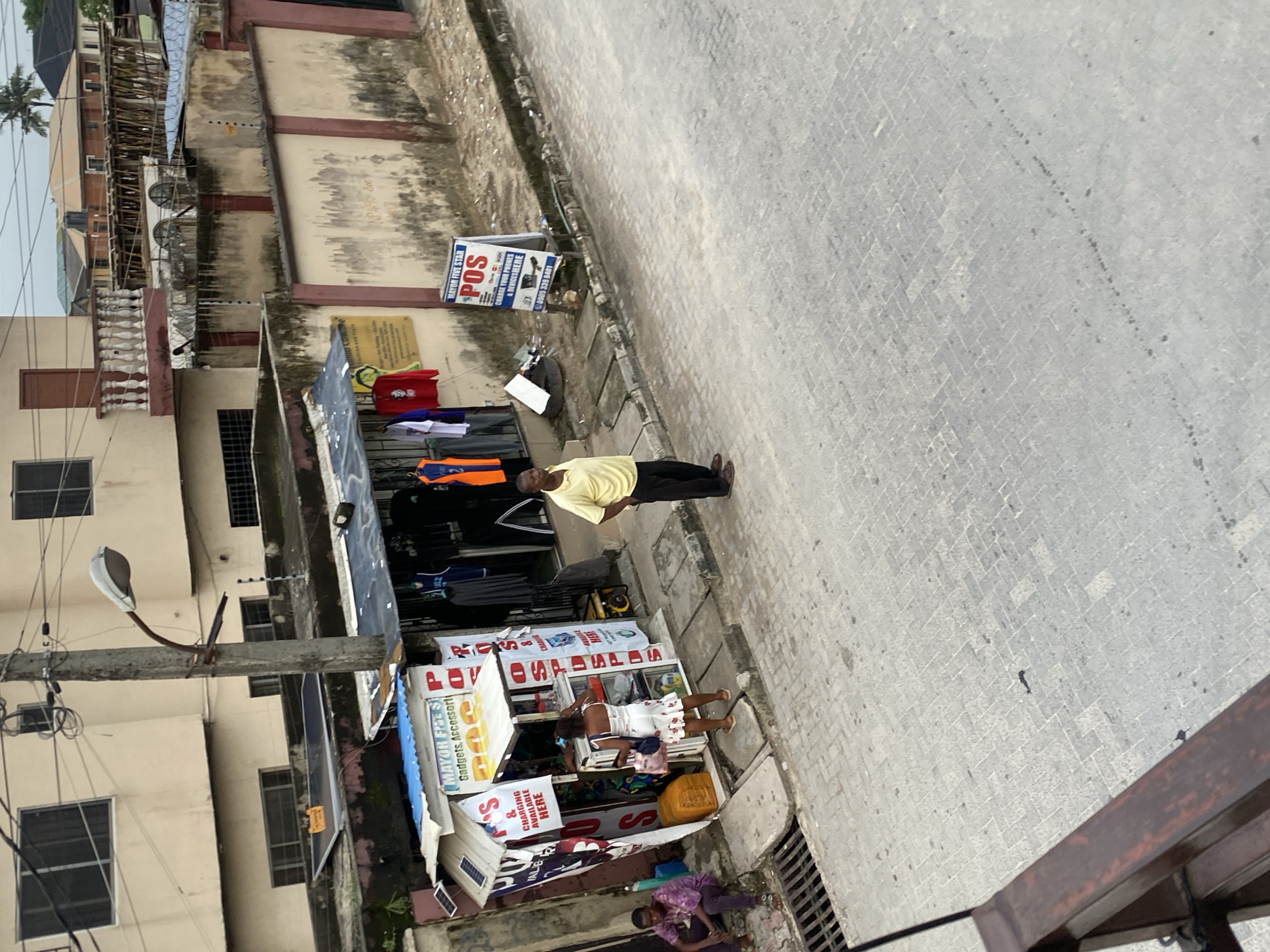 An urban street scene in Ado, Nigeria, shows a man in a yellow shirt standing on a paved sidewalk next to several small storefronts. The storefronts sell phone chargers and gadgets. One sign advertises "MAYOR FIVE STAR POS" and indicates phone charging services are available. Another sign reads "CHARGING AVAILABLE HERE". A woman in a white floral dress is visible within one of the storefronts. Clothing hangs outside another store. A concrete building lines the back of the sidewalk, and a light pole is on the left side. A person is seated on the ground near a storm drain.