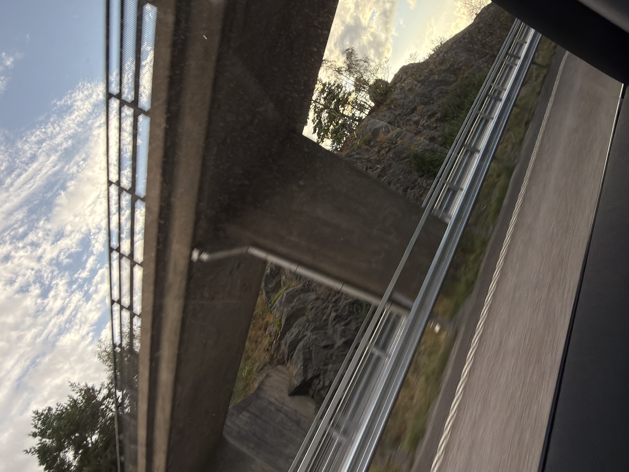 The image shows a view from a vehicle moving along a road, likely a highway, in Oskarsberg, Sweden. The perspective is from inside the vehicle, looking out and slightly upwards towards an overpass or bridge.
The overpass is constructed of concrete, with a solid lower section and a railing along the top. The railing has a distinctive pattern of rectangular openings, filled with a transparent or reflective material. Through the openings, a cloudy sky is visible, with a hint of trees and rocky terrain.
Beneath the overpass, a steep, rocky incline is visible, sparsely covered with vegetation. The road in the foreground is paved and has lane markings. A metal guardrail runs along the side of the road.
The lighting suggests it is either early morning or late afternoon, with the sun casting a warm glow on the sky and landscape. There are no people visible in the image. The overall impression is of a typical Swedish roadside landscape with infrastructure.