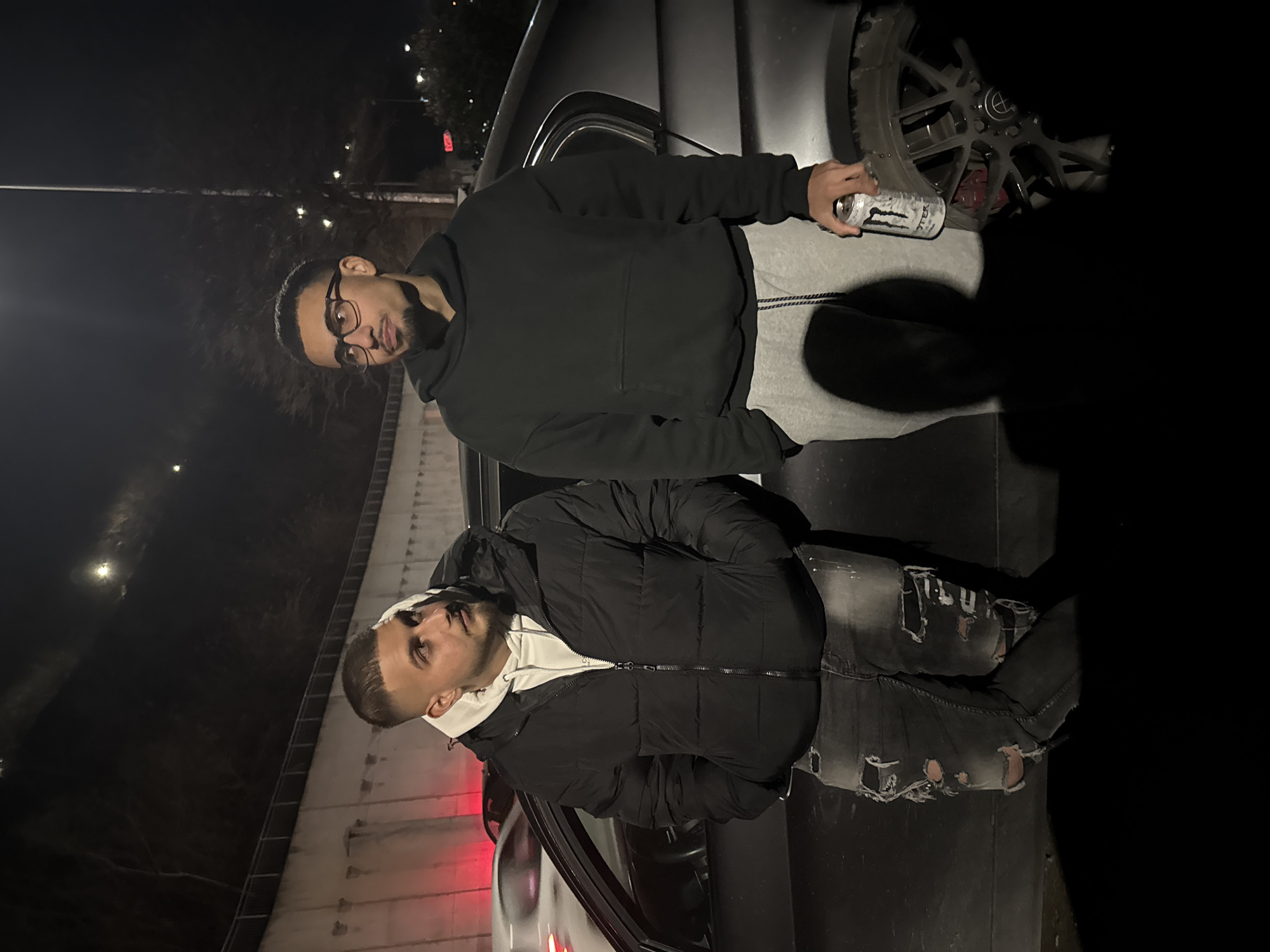 Two men are posing in front of a car. The man on the top is wearing a black hoodie, grey pants, and glasses and is holding a can. The man on the bottom is wearing a black puffer jacket over a white hoodie and ripped jeans. The car is silver and the location appears to be at night in an urban setting. The location can't be verified as the city of Varna, Bulgaria without additional verifiable evidence.