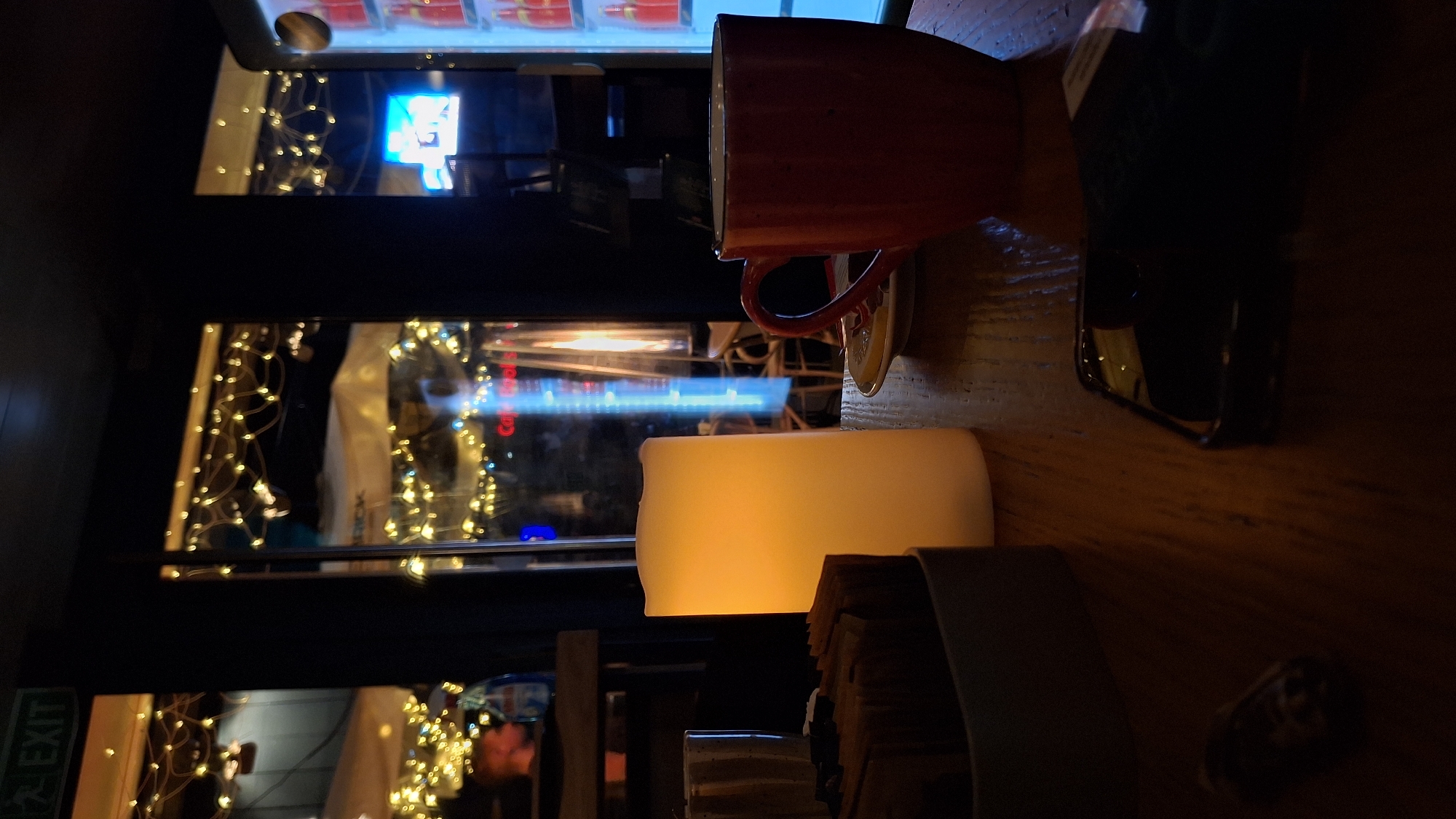 A rotated interior shot shows a cafe table with a red mug on a saucer, a lit faux candle, a stack of coasters, a mobile phone, and a bottle cap. The table surface has a wood grain pattern. In the background, windows are adorned with small lights, and a sign reading "EXIT" is visible. The cafe is located in Varna, Bulgaria, as evidenced by the reflection in the window of street signage in Cyrillic script. No people are clearly visible.