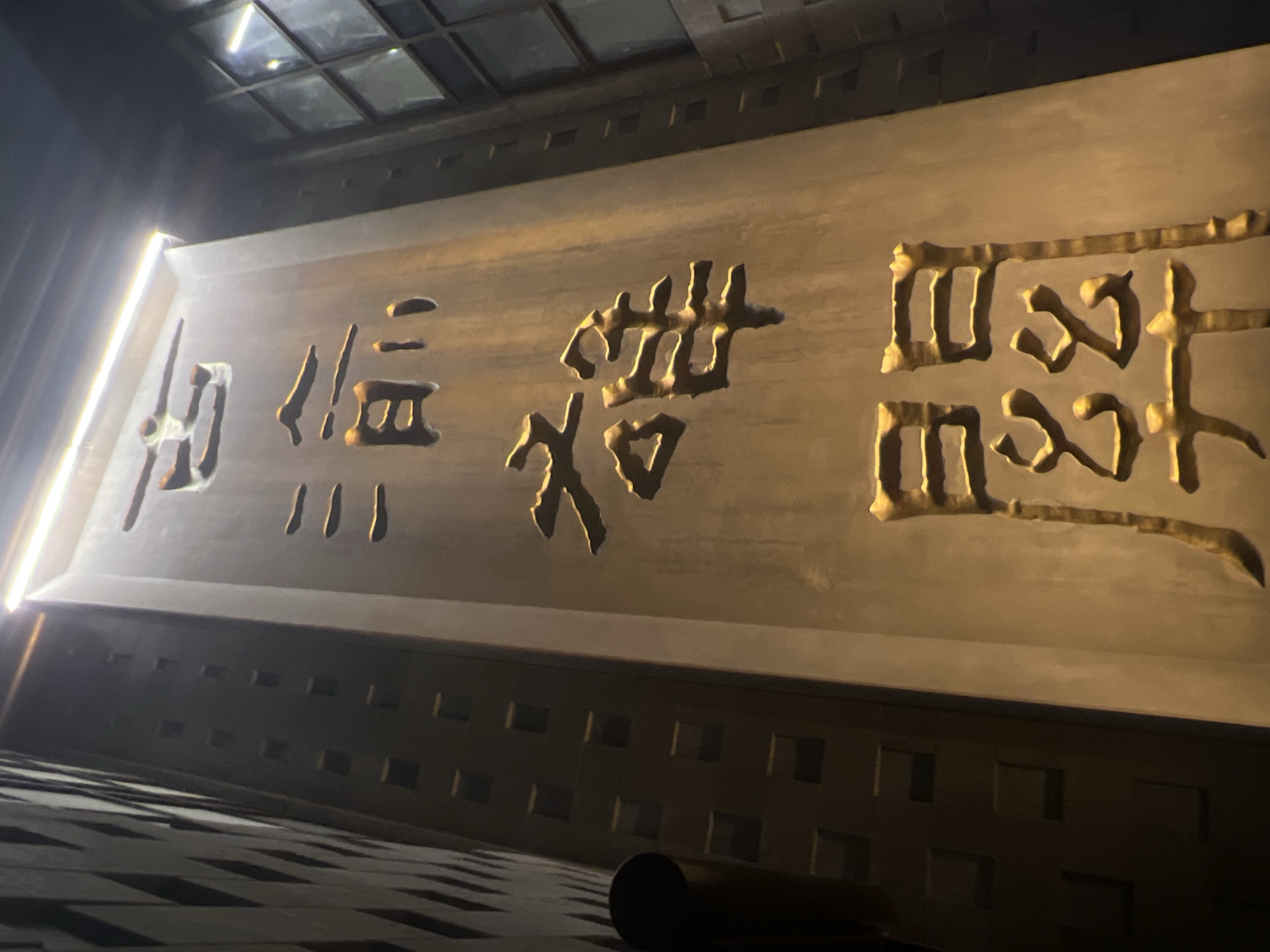 The image shows a close-up, low-angle view of a textured, vertical monument illuminated by a bright light at the top. The monument is made of a material that appears to be stone or metal, with Chinese characters carved into its surface. From top to bottom, the characters read: "古 渝 雄 關". The first character, "古" (gǔ), meaning "ancient," is at the top. Below it is "渝" (yú), the abbreviation for Chongqing. Next is "雄" (xióng), meaning "heroic" or "grand." Finally, at the bottom is "關" (guān), meaning "pass" or "gate." Together, the characters can be translated as "Ancient Chongqing Pass" or "Grand Pass of Old Chongqing."
The monument is positioned in front of a modern building with a patterned facade, characterized by a diamond-like texture. To the right of the monument, a windowed section of another building is visible, its panes reflecting the ambient light. The overall scene suggests an outdoor setting in a city, likely a historical or landmark area. The lighting creates strong shadows and highlights, emphasizing the depth and texture of the monument and its inscriptions. The image quality is slightly grainy, particularly in the darker areas. The context provided mentions Yuzhong District, China, which is a central district of Chongqing. This monument is likely a notable landmark within that district.