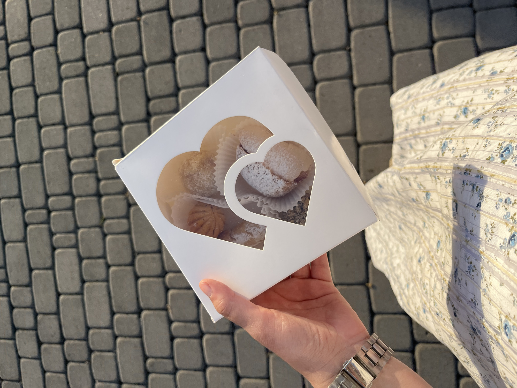 The image shows a person's hand holding a white cardboard box containing several pastries. The box has heart-shaped cutouts revealing the treats inside, which appear to be small, round cakes dusted with powdered sugar and possibly filled with jam or fruit. The pastries are arranged in white paper liners.

The person is wearing a silver wristwatch with a metal band. Their arm is draped over a white garment with a delicate blue floral pattern, suggesting a dress or blouse. The background consists of grey paving stones laid in a repeating pattern, indicating an outdoor setting.

The lighting suggests it is daytime, with sunlight casting shadows on the paving stones. The overall mood seems pleasant and casual, perhaps a moment of enjoying a treat outdoors. The location context provided is Dubivtsi, Ukraine. No visible text is present in the image.