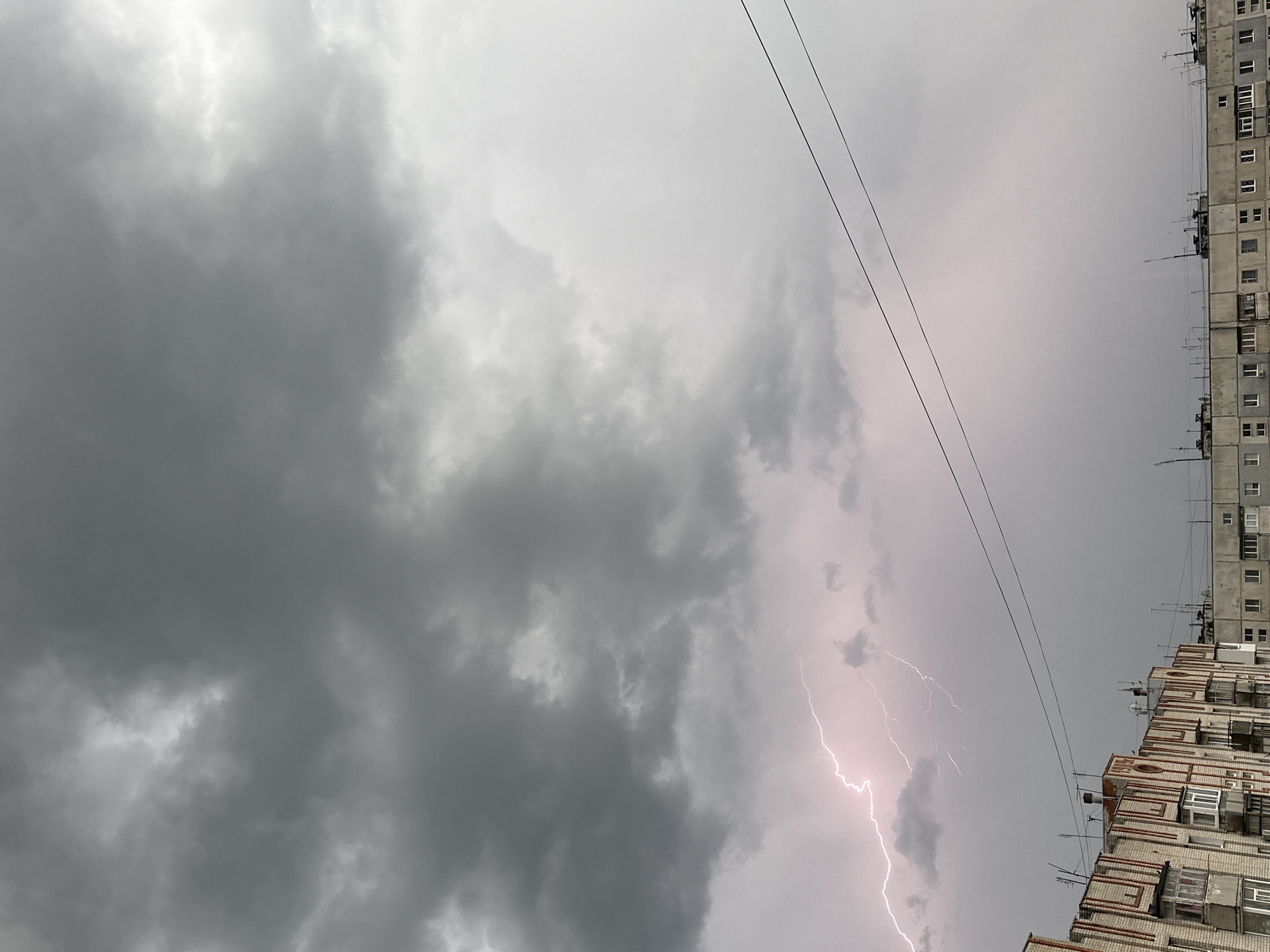 The image shows a cityscape during a thunderstorm in an unknown city. Dark, heavy clouds dominate the sky, with a prominent lightning strike visible in the right-center of the frame. A tall, multi-story apartment building is partially visible on the right side, and power lines stretch across the upper portion of the sky. The overall environment suggests inclement weather and a tense atmosphere.
