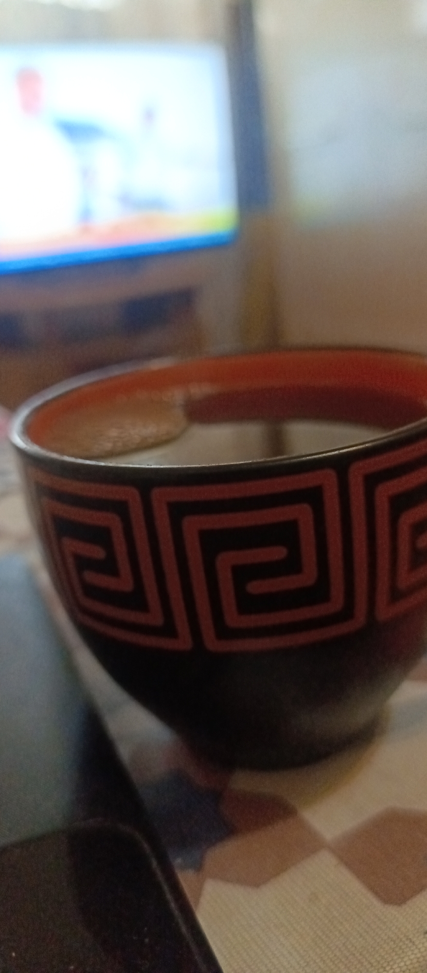 The image shows a close-up of a black and red patterned bowl filled with a dark liquid, likely tea or coffee. The bowl is resting on a patterned surface, possibly a tablecloth. In the blurred background, a television screen is visible, displaying a bright and colorful image. The scene appears to be indoors, in a casual setting. There are no people visible in the image. The location in the city of Voskresenskiy, Russia, is not discernible from the image content.