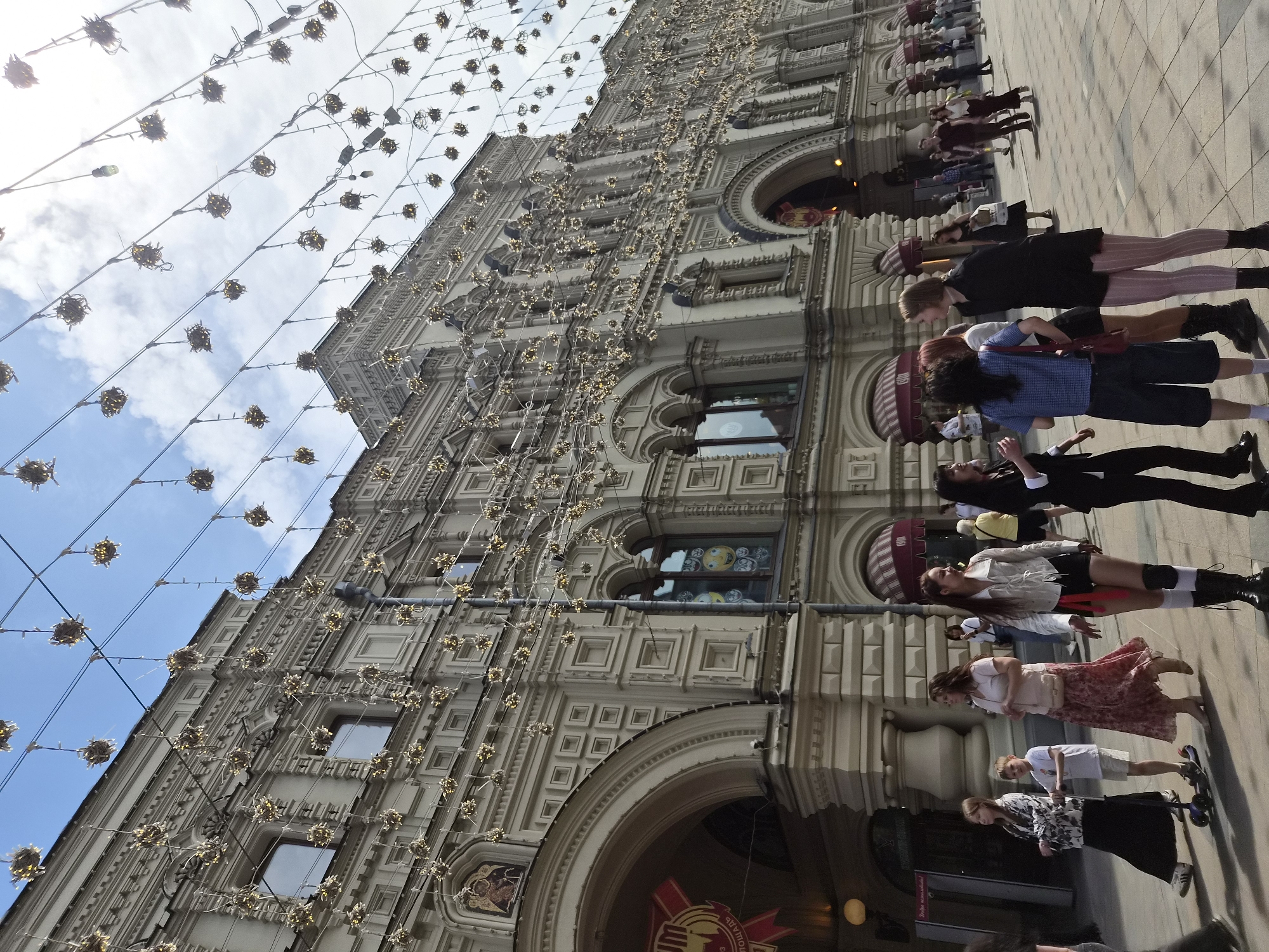 This image depicts the facade of the State Department Store (GUM) on Red Square in Moscow, Russia. The multi-story building is constructed in a light-colored material, featuring an ornate architectural style with numerous decorative elements including cornices, pilasters, square relief panels, and arched windows and doorways. An arched entrance on the left side displays a partially visible red and white banner with text and the year "1893". Above this arch, a religious mosaic icon is integrated into the facade. Another window to the right shows reflections of yellow smiley faces. A grey downspout runs vertically along the facade near the center-right.
Overhead, numerous strings of decorative lights are suspended across the visible sky. These lights consist of small, spherical, starburst-like clusters emitting a golden glow, creating an illuminated canopy. The sky above is partially blue with scattered white clouds.
On the paved street level in front of the building, a diverse group of people is present. In the foreground, from left to right: a young girl in a patterned long-sleeved top and dark skirt walks alongside a young boy riding a blue scooter. Adjacent to them, a woman in a light pink blouse and a long floral skirt walks to the right. To her right, another woman is depicted wearing a light-colored jacket, dark shorts, white knee-high socks, and black boots, with red fabric strips hanging from her waist. Further right, a woman in a black dress and pink vertically striped tights stands, conversing with others, including a woman in a blue checkered shirt and dark shorts. In the mid-ground and background, additional individuals and groups of people are seen walking and standing on the tiled pavement. Shadows are cast on the ground, indicating direct sunlight from a high angle.