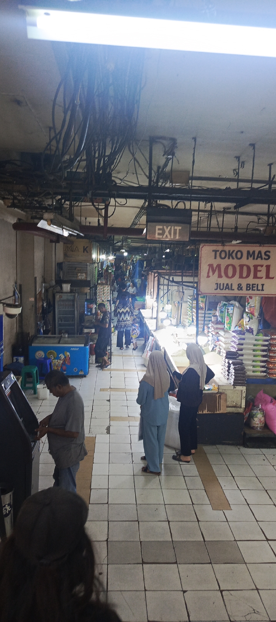 The image depicts an indoor marketplace scene. A fluorescent light fixture is visible at the top, with a cluster of black electrical cables hanging from the ceiling. An "EXIT" sign is mounted above a doorway. To the right, a sign reads "TOKO MAS MODEL JUAL & BELI," indicating a gold shop that buys and sells. Several people are present, including a man in a grey shirt using an ATM machine on the left, and three individuals in the middle wearing headscarves, two of whom are facing away from the camera. Further down the aisle, more people are visible amidst market stalls stocked with various goods, including what appear to be bags of rice. A blue refrigerated display case is also present. The floor is tiled with a grid pattern. The overall environment suggests a busy, somewhat cluttered commercial space. The presence of the "TOKO MAS MODEL" sign strongly suggests the location is in Indonesia, and the city of Sukaresmi is a district within the Cikole subdistrict of the city of Sukabumi in West Java, Indonesia.
