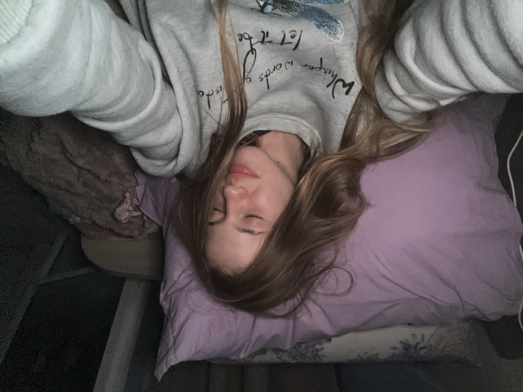 A young woman with long blonde hair is lying in bed, eyes closed, with a peaceful expression. She is wearing a light gray sweatshirt with a design and text on it. The sweatshirt has a ribbed texture around the cuffs and neckline. She is lying on a lavender pillow, and other bedding and pillows are visible in the background, suggesting a bedroom setting. The lighting is dim, indicating it may be nighttime or a dimly lit room. The overall scene evokes a sense of rest and comfort. The location in the city of Unknown, Russia is not discernible from the image.