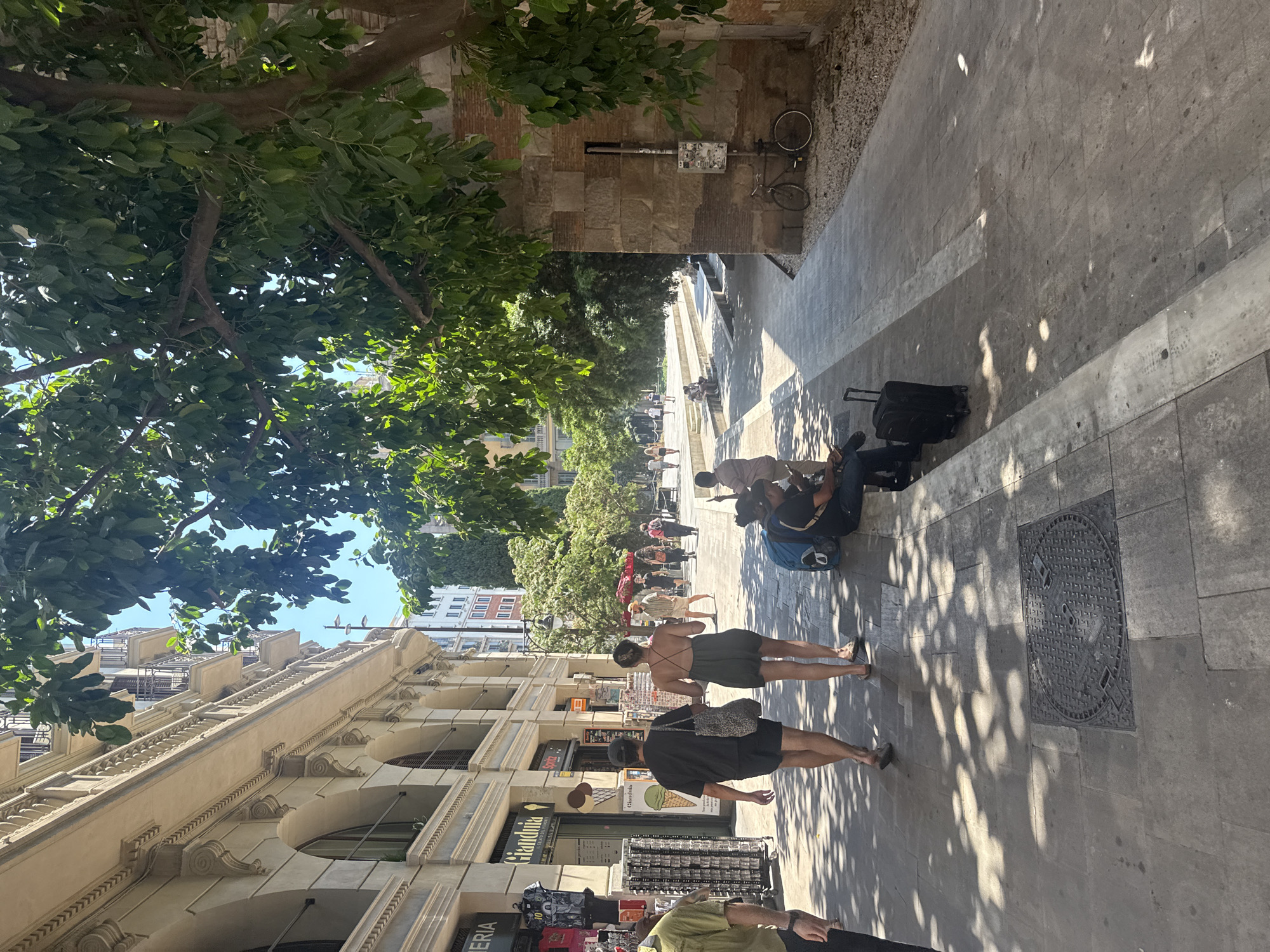 This image depicts a street scene in Barcelona, Spain on a bright, sunny day, likely in the late morning or early afternoon given the strong shadows. The perspective is from a high angle, looking down at a pedestrianized area adjacent to a building with classic European architecture.
The foreground is dominated by lush green tree foliage, partially obscuring the view and casting dappled shadows on the ground. Below, several people are visible walking and standing. A woman in a dark green dress and another in a black t-shirt and shorts are walking away from the camera. Further down the street, a group of people are seated near a water feature or fountain, enjoying the sunshine. Luggage, including a black suitcase, suggests travel or movement. In the background, more trees and buildings line the street.
A bicycle is parked against a textured brick wall in the upper right portion of the image. The building to the left displays ornate architectural details, including balconies with decorative railings and arched window frames.
Some text is visible on the storefronts. On the left, partially obscured, "Gelateria" is visible. Below it, "Gianduja" and "Cioccolato" are clearly legible, suggesting an ice cream shop or confectionary. Another sign reads "Sarra". The overall impression is a vibrant and lively urban environment on a pleasant day.