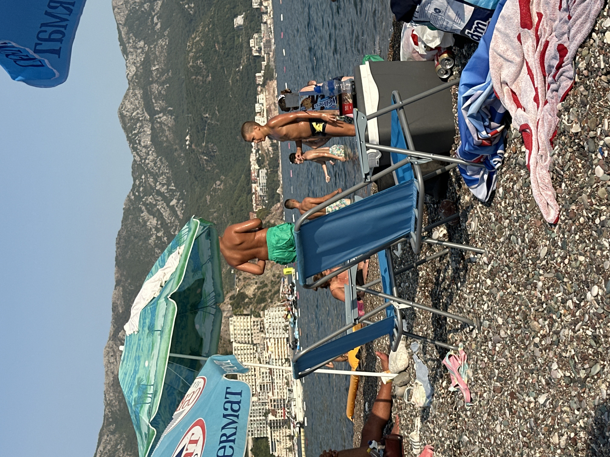 The image depicts a beach scene in Boreti, Montenegro, under a clear sky. In the foreground, several beach chairs and towels are arranged on a pebbled shore. A few people are visible in the water, with mountains and buildings forming the backdrop. The water appears calm, and the overall atmosphere suggests a typical summer day at the beach.