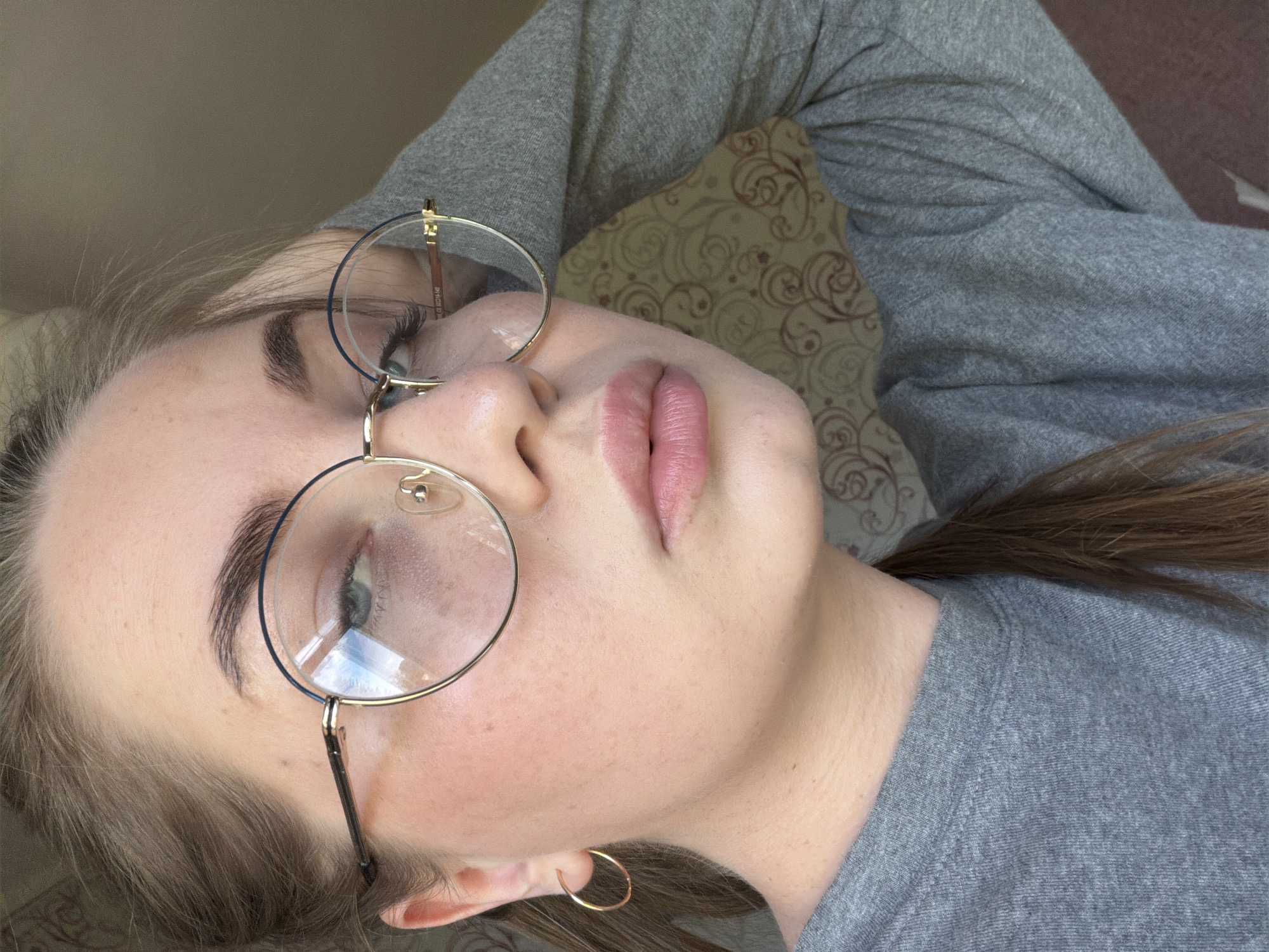 A person with light skin and blonde hair is lying down, facing upwards. The person is wearing round glasses with gold frames. The person is wearing a gray long-sleeved shirt. The background includes a pillow with a brown patterned design. The person is in Уральское, Russia.