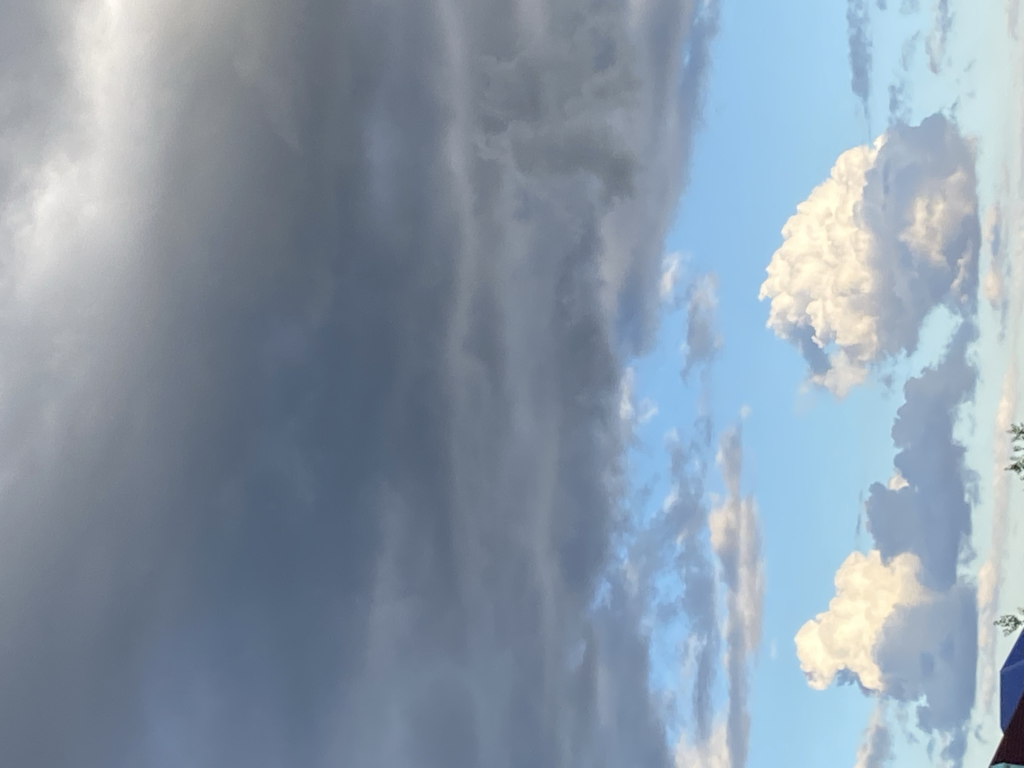 The image depicts a sky with layered clouds. The upper portion of the sky is filled with dark, heavy storm clouds, creating a somber atmosphere. Below these, a band of lighter, wispy clouds stretches across a bright blue sky. At the bottom of the frame, two large, fluffy cumulus clouds are visible, illuminated by the sun. A sliver of a red and blue roofline and some greenery are visible at the very bottom edge of the image, suggesting a terrestrial viewpoint. The scene is characteristic of the sky over Байқоныс ауылдық округі, Kazakhstan, where such dramatic cloud formations are common. There are no people or specific objects visible that would indicate an action or interaction. The environment is solely focused on the sky and its atmospheric conditions.