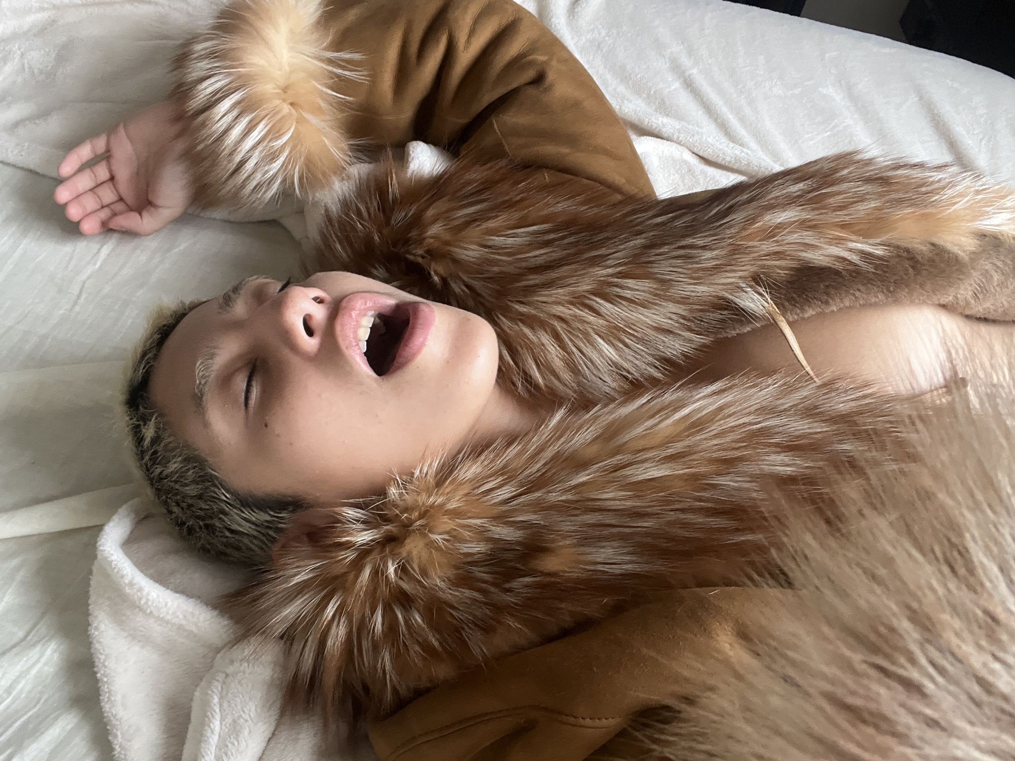 The image shows a person lying on a white bed or blanket. The person has fair skin, short, dyed hair with darker roots, and their mouth is open. They are wearing a brown jacket with a fur collar. The hand is visible on the left side of the frame. The background is a plain white surface. There is no indication of the location being in Buenos Aires, Argentina.