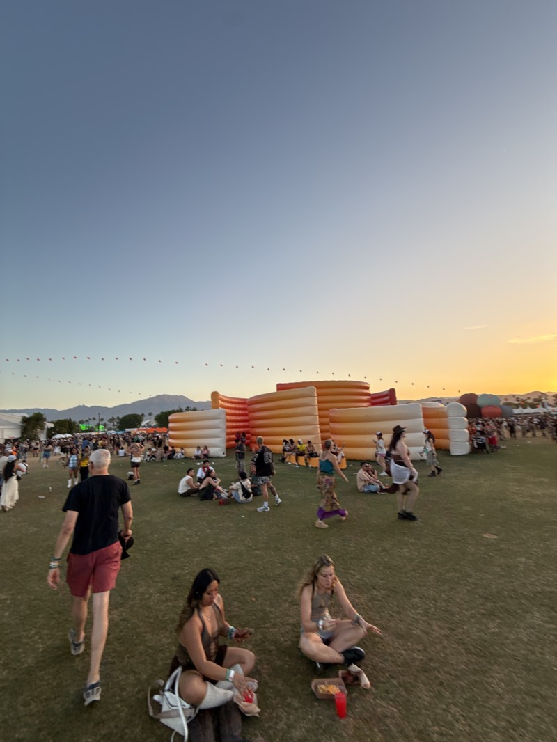 The image captures an outdoor festival or large gathering set on a sprawling green grassy field in Indio, United States, during the late afternoon or early evening. The sky displays a clear gradient from deep blue overhead to a warm orange and yellow near the horizon, signifying a sunset. Distant mountains are faintly visible along the skyline.
Numerous attendees populate the field. In the foreground, two women are seated cross-legged on the grass; one, wearing a brown ensemble, has a white bag beside her, while the other, in a light top, has a red cup and a small food container. A man in a black t-shirt and red shorts walks away from the viewer in the lower-left. Further back, many groups of people are sitting, standing, and mingling across the expansive lawn.
A large, multi-tiered inflatable structure, composed of interconnected orange and white sections, dominates the mid-ground, serving as a focal point or a communal resting area. A string of lights or small red objects can be seen stretching horizontally across the distant horizon, adding to the festival's illuminated atmosphere. The overall scene conveys a relaxed and social environment, with people enjoying the outdoor setting and event. There is no clearly legible text visible within the image.