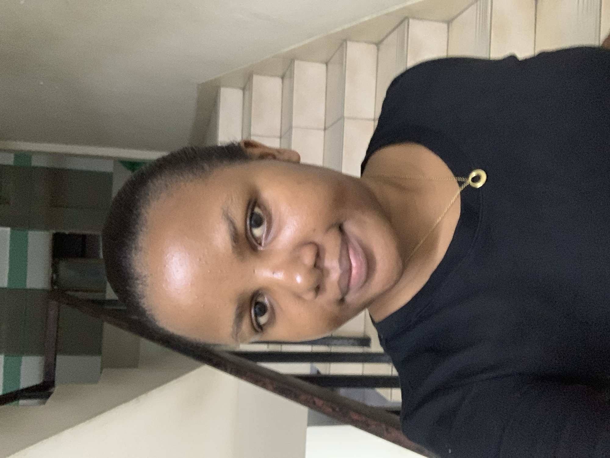 A young, dark-skinned woman wearing a black top and a gold necklace is indoors. The background features a staircase with off-white tiled steps. A railing with black bars is also visible. The location appears to be a residential building. It is not possible to determine the city of Dar es-Salaam, Tanzania from the given image.