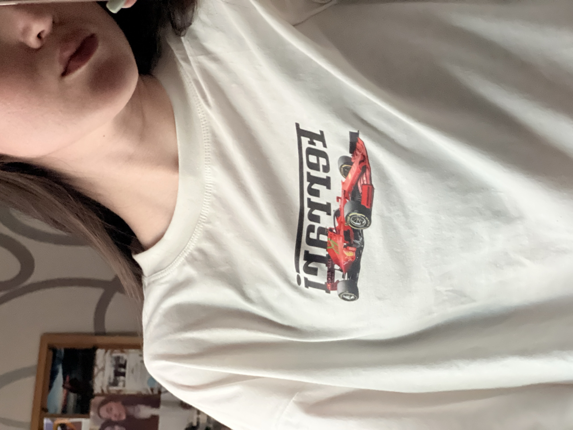 A close-up, waist-up shot shows a person wearing a white t-shirt with a graphic of a red Formula 1 car and the text "1991" in black. The person's face is partially visible, with their mouth and chin in focus. The background is a wall with a framed collage of pictures and a faint circular pattern. The lighting suggests an indoor setting, possibly a bedroom. There are no discernable elements to indicate a location in Kazan, Russia.