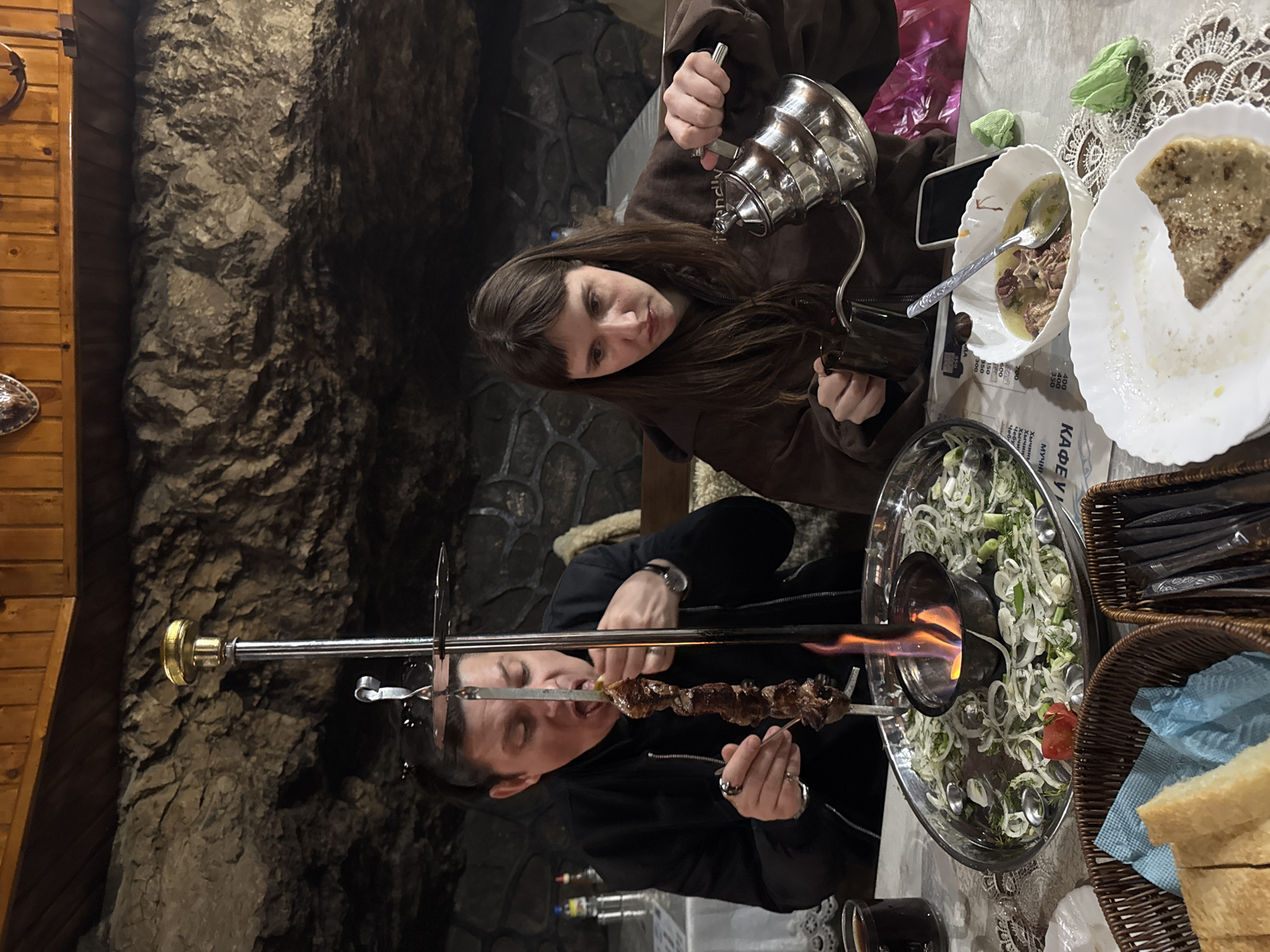The image captures a dining scene featuring two individuals in a rustic restaurant setting, likely in Khushto-Syrt, Russia. The interior features dark, rough stone walls, giving a cave-like ambiance, alongside wooden paneling.
In the foreground, a man wearing a black jacket is engaged in eating. He holds a skewer of grilled meat, such as shashlik, which is positioned over a small fire pit at the center of a large, circular metal platter. This platter also holds a vibrant salad of sliced onions, herbs, and tomatoes, suggesting a communal and interactive dining experience where meat is kept warm over the flame.
To the right, a woman with long dark hair, dressed in a brown hoodie, is pouring liquid from a silver metal kettle into a cup. On the table, there are white plates; one holds a piece of flatbread, and another contains a bowl of soup or stew. A basket with bread and blue napkins, and a smartphone are also visible on the table.
The lighting is warm and artificial, creating an intimate atmosphere that accentuates the flame under the skewers. Visible text on what appears to be a menu or newspaper includes Cyrillic words such as "КАФЕ У" (Cafe at/near), "МУЧНИ" (flour products), "Хычины" (Khychiny – a traditional flatbread), and "Чебу" (Cheburek – another pastry), accompanied by numbers that are likely prices (e.g., 600, 400, 350). The scene conveys a relaxed and enjoyable meal, focusing on traditional regional cuisine.