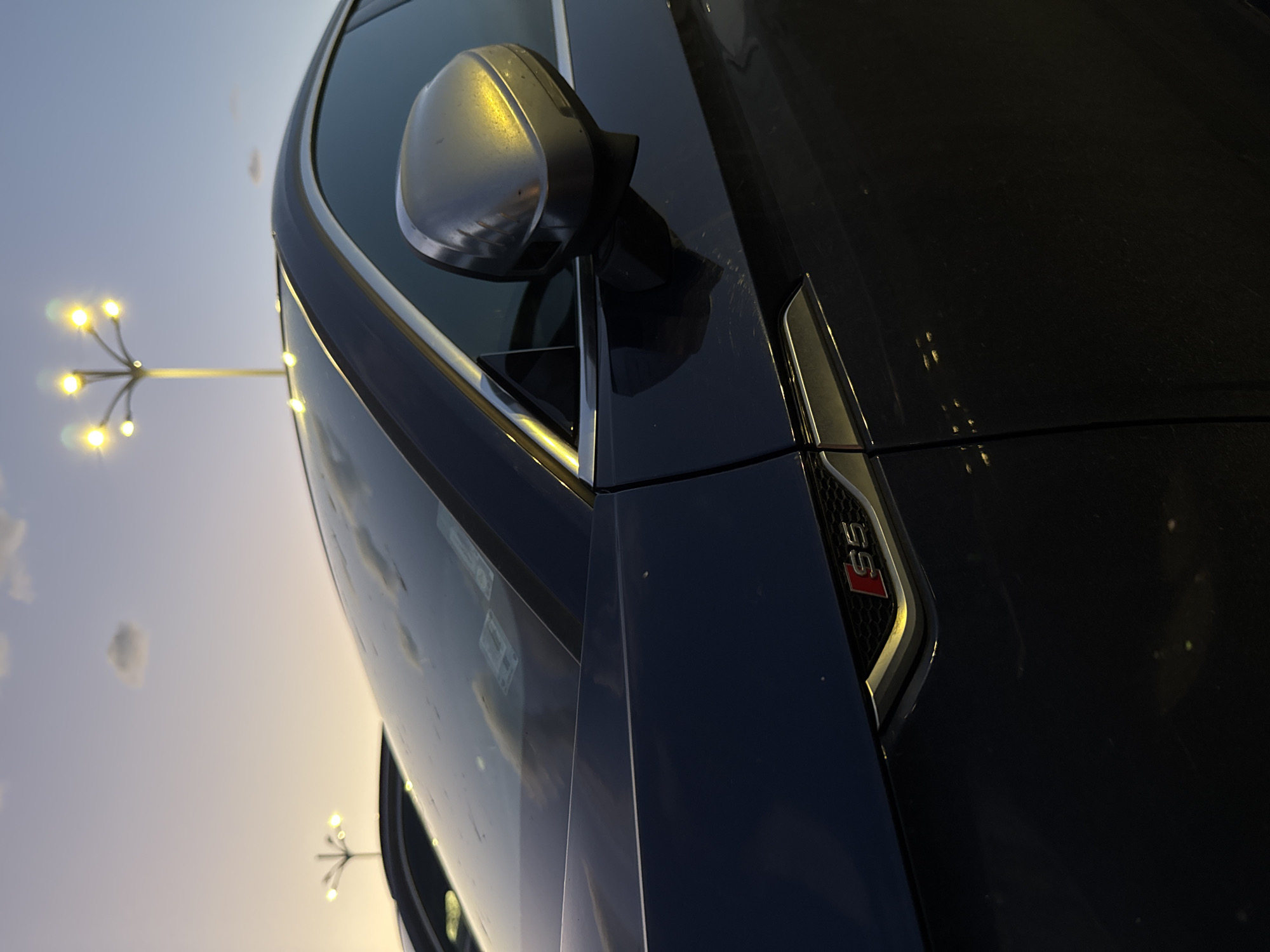 The image shows a close-up view of the front quarter panel of a dark-colored car, specifically an Audi S5, as evidenced by the S5 badge. The car is parked outdoors with a street lamp visible in the background. The location is Plovdiv, Bulgaria, though no specific landmarks are apparent in the image. The focus is primarily on the car's design details, including the side mirror and the body lines. The environment suggests it is either dusk or dawn, due to the soft lighting conditions. No people are visible.