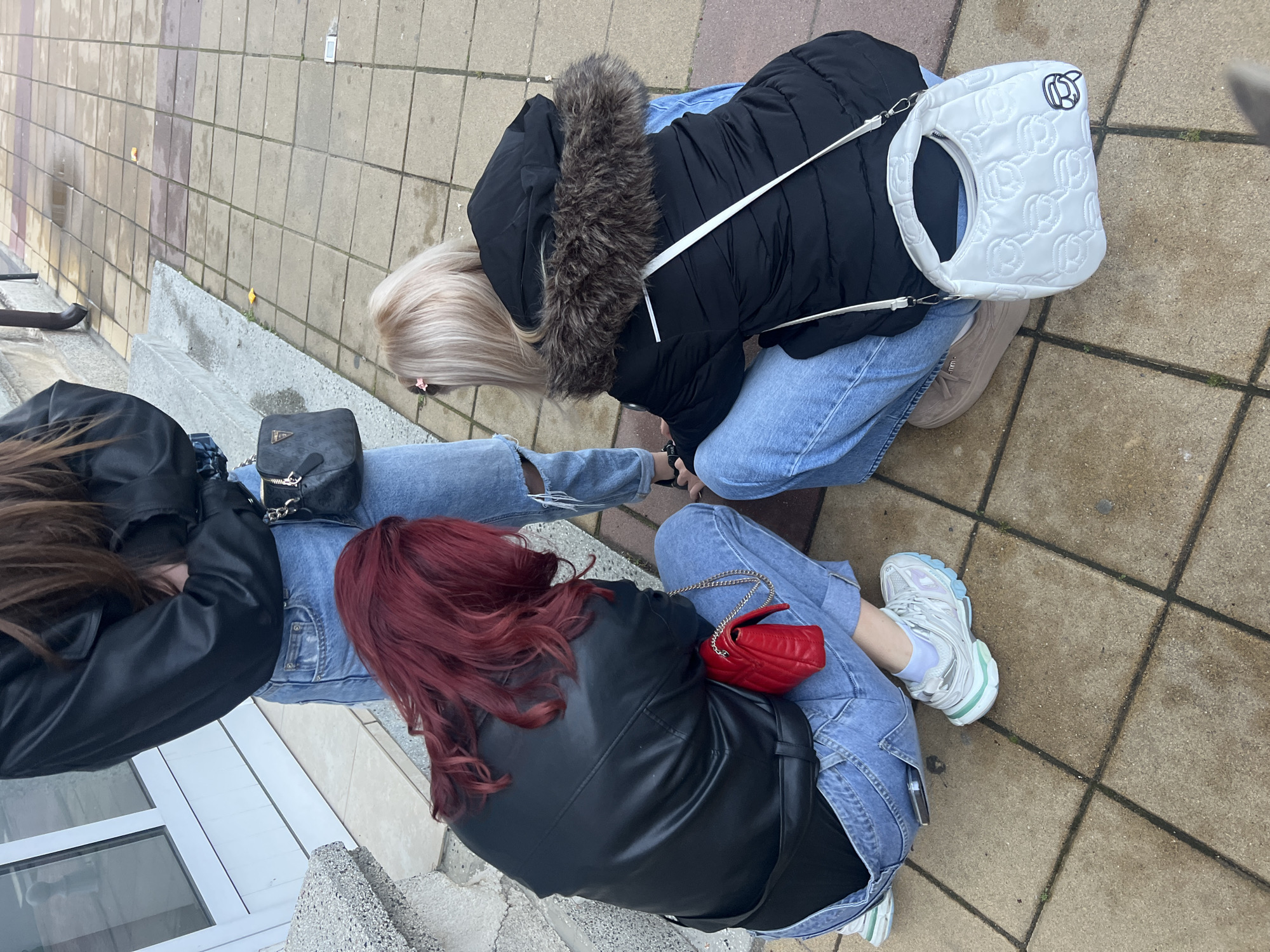 Three people are sitting on a ledge in the city of Unknown, Unknown. The person on the left has long brown hair, and they are wearing a black leather jacket and blue jeans. A black purse is resting on their lap. To the right of this person, another person with red hair is wearing a black leather jacket and blue jeans, with a small red purse on their lap. They are wearing white sneakers with a hint of pale green on the soles. The third person, on the right, has blonde hair and is wearing a black puffer jacket with a fur-lined hood and blue jeans. A white shoulder bag is next to them. The background consists of paving stones and a white wall.