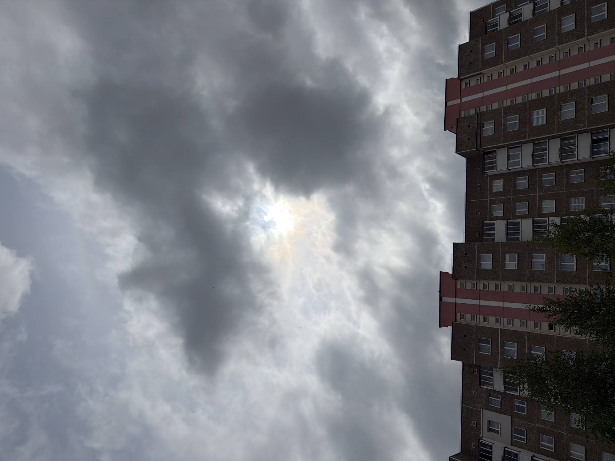 The image depicts a tall, likely residential building in an unknown city. The building has a repetitive pattern of windows and distinct horizontal red stripes. The sky is heavily overcast with a break in the clouds revealing the sun. Part of a tree is seen in the right side of the image.
