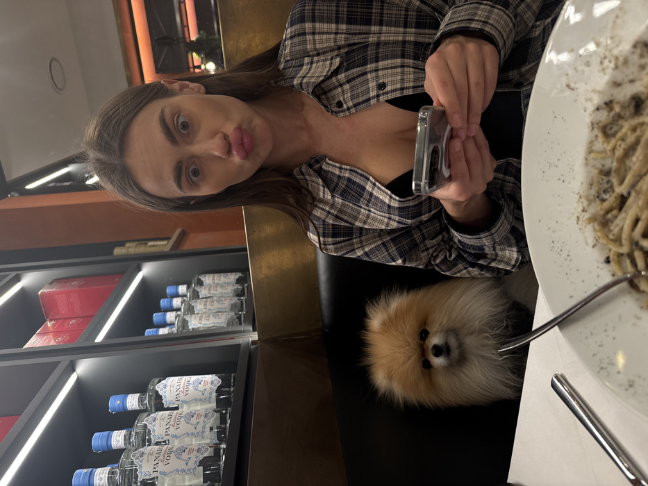 The image depicts an indoor dining establishment in Verona, Italy. A young adult female is seated at a table to the right, posing with wide eyes and pursed lips. She has long brown hair and is wearing a blue and white plaid shirt over a black top. She holds a smartphone with a clear case in both hands at chest level. To her left, a small, fluffy, tan-colored Pomeranian dog is seated on the dark upholstered bench, looking directly at the camera. In the foreground, a white tablecloth is visible with a white plate containing partially consumed pasta, likely with truffle or pepper, and metal cutlery. The background features dark shelving units with integrated strip lighting, displaying multiple bottles of "PANAREA VODKA" with blue caps and white labels, along with some red boxes. A warm-toned panel or wall runs beneath the shelves.