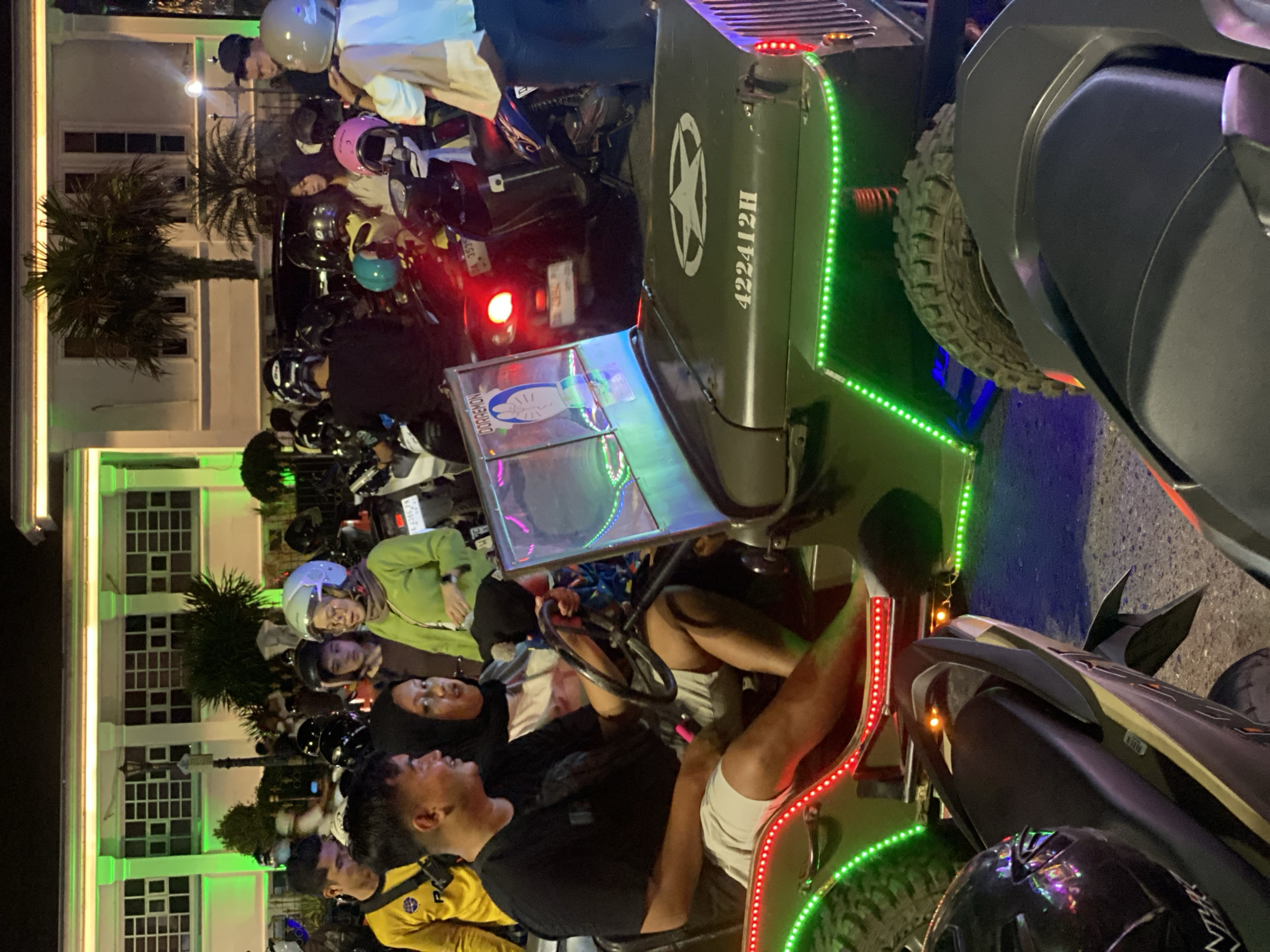A group of people are gathered around a vintage military-style jeep adorned with colorful LED lights, parked in front of a building with green exterior lighting. The scene appears to be a street at night, possibly in the city of Banjarbaru, Indonesia, given the presence of motorcycles and a bustling atmosphere.
In the foreground, a man and a woman are seated in the jeep, with the woman holding the steering wheel. The man is wearing a black t-shirt and light-colored shorts, while the woman has a headscarf and a light-colored top. Behind them, other individuals are standing, some wearing helmets, suggesting they arrived on motorcycles. The jeep itself is olive green and features a white star emblem on its hood, along with the number "422412H". The vehicle is decorated with green and red LED strip lights along its sides, illuminating the surrounding area.
In the background, several motorcycles are parked. A building with large windows and a white facade is visible, illuminated by bright green lights along its top edge. Palm trees are present in the background, adding to the tropical ambiance. The overall impression is a lively evening gathering, likely a social event or a street fair.