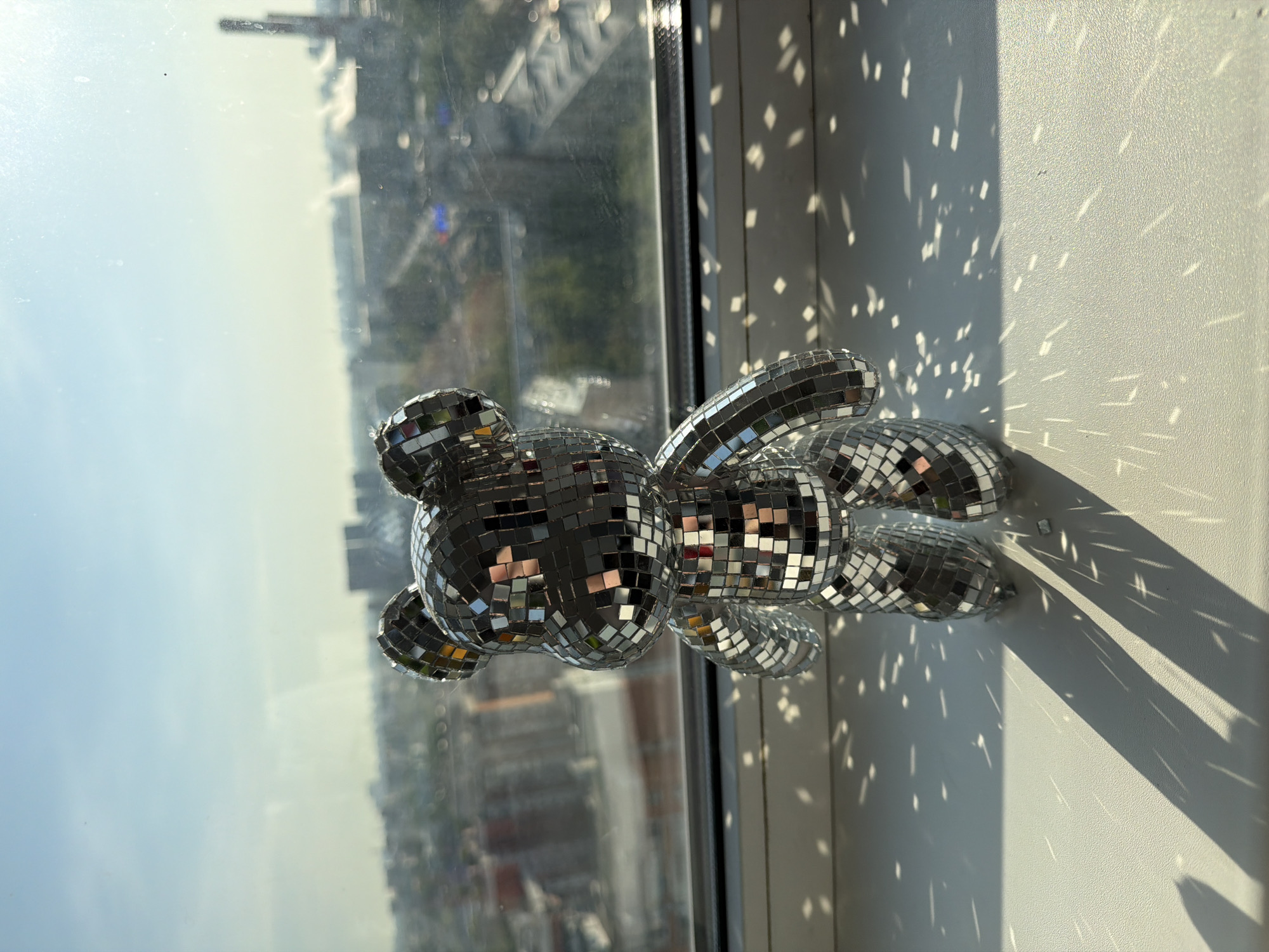 A disco ball teddy bear lies on a window sill in an unknown city. The light scatters on the wall next to the window, creating patterns of light and shadow. The window looks out onto a hazy cityscape.