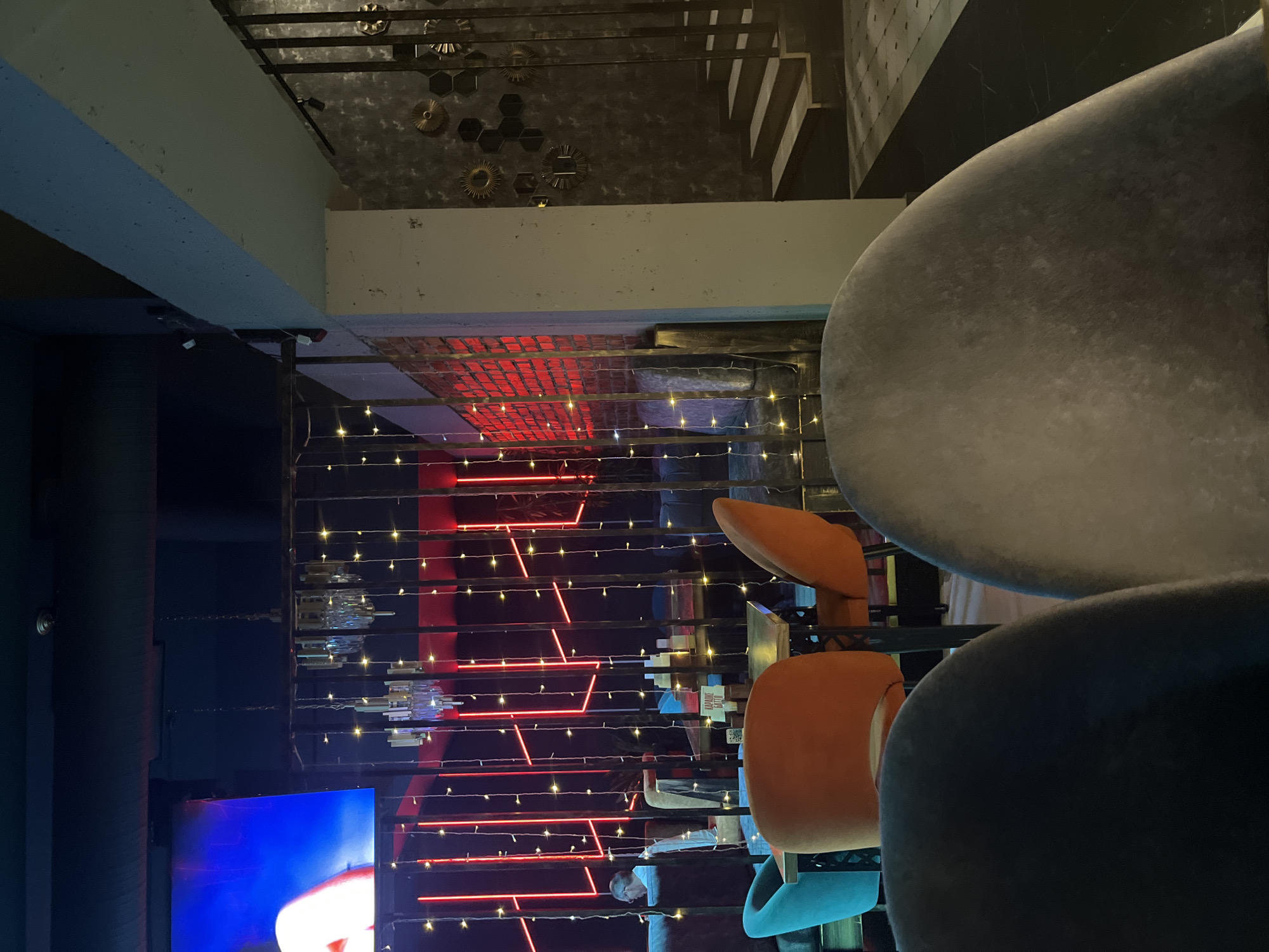 The image depicts the interior of a dimly lit establishment, possibly a cafe or restaurant, with a focus on seating areas. In the foreground, two chairs with plush, grey upholstery are prominently featured. One chair is dark grey and the other is a lighter, textured grey. Behind these chairs, several other seating arrangements are visible, including a bright orange chair and a teal chair, both with backs. These chairs surround a dark-colored table.
Further back, a series of vertical metal bars creates a partition, behind which red neon lights form geometric patterns against a brick wall. Small, twinkling fairy lights are strung across the bars, adding a decorative element. A glimpse of a television screen displaying a blue and pink image is visible in the upper left portion of the frame. A person wearing a white shirt is seated at a table in the background, partially obscured by the metal bars.
To the right of the metal partition, a dark wall with decorative metallic circular and hexagonal shapes is visible. A staircase with dark steps and a black railing ascends to the upper level. The floor in this area appears to be tiled with a checkerboard pattern of dark and light squares. The overall atmosphere suggests a modern, stylish, and possibly entertainment-oriented venue.
This establishment is identified as "Karaoke Bar" based on a sign on a table. The scene is likely within the city of Stavropol, Russia, given the context provided.