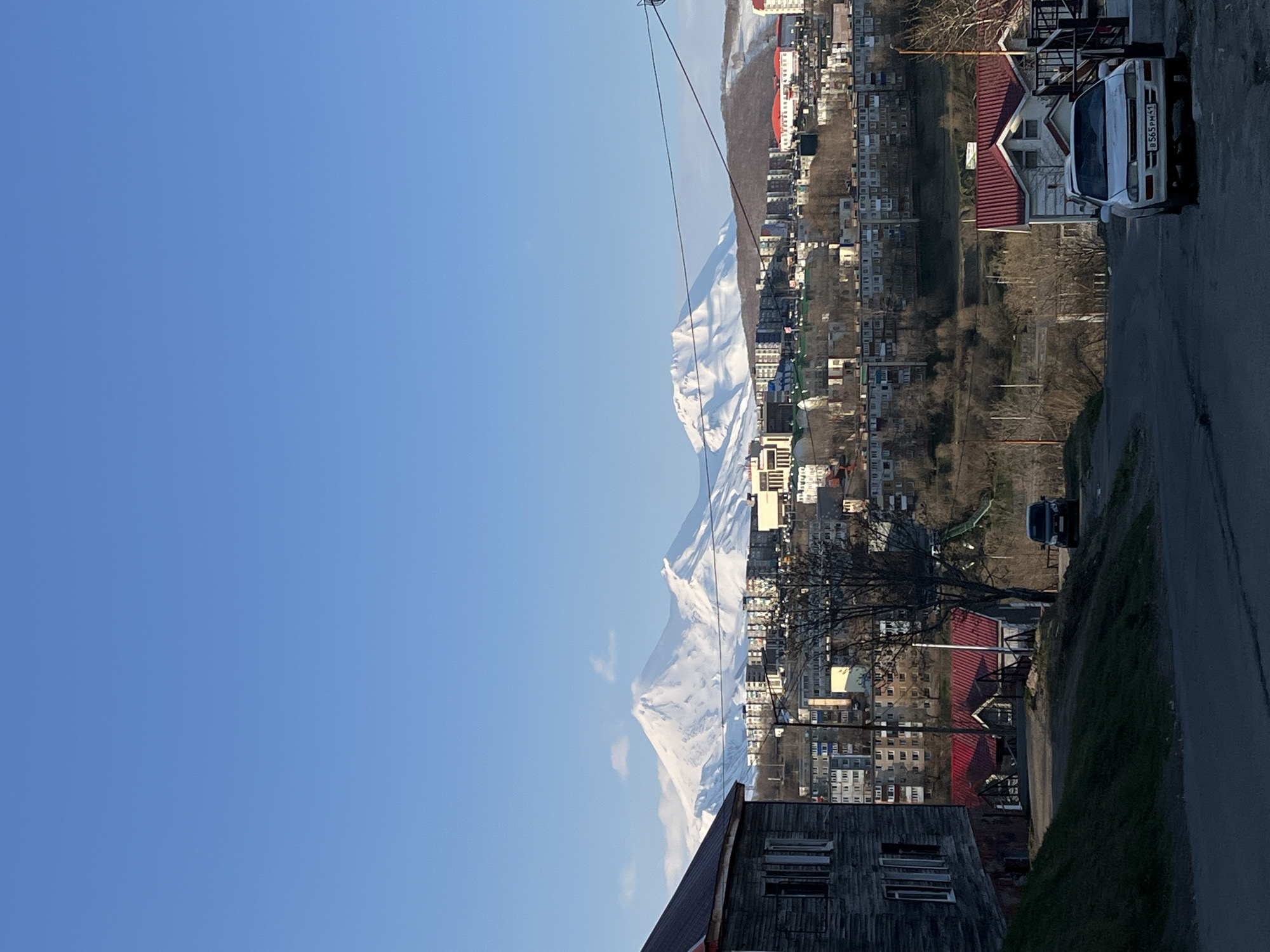 The image depicts a cityscape of Petropavlovsk-Kamchatsky, Russia, featuring a clear blue sky. Visible are buildings and dwellings of varying sizes and roof colors (predominantly red). Mountains are partly covered in snow, and utility lines can be seen. A road is visible, with a white van parked on the side. Some vegetation is present, including trees without leaves. The perspective suggests the photo was taken from an elevated position.