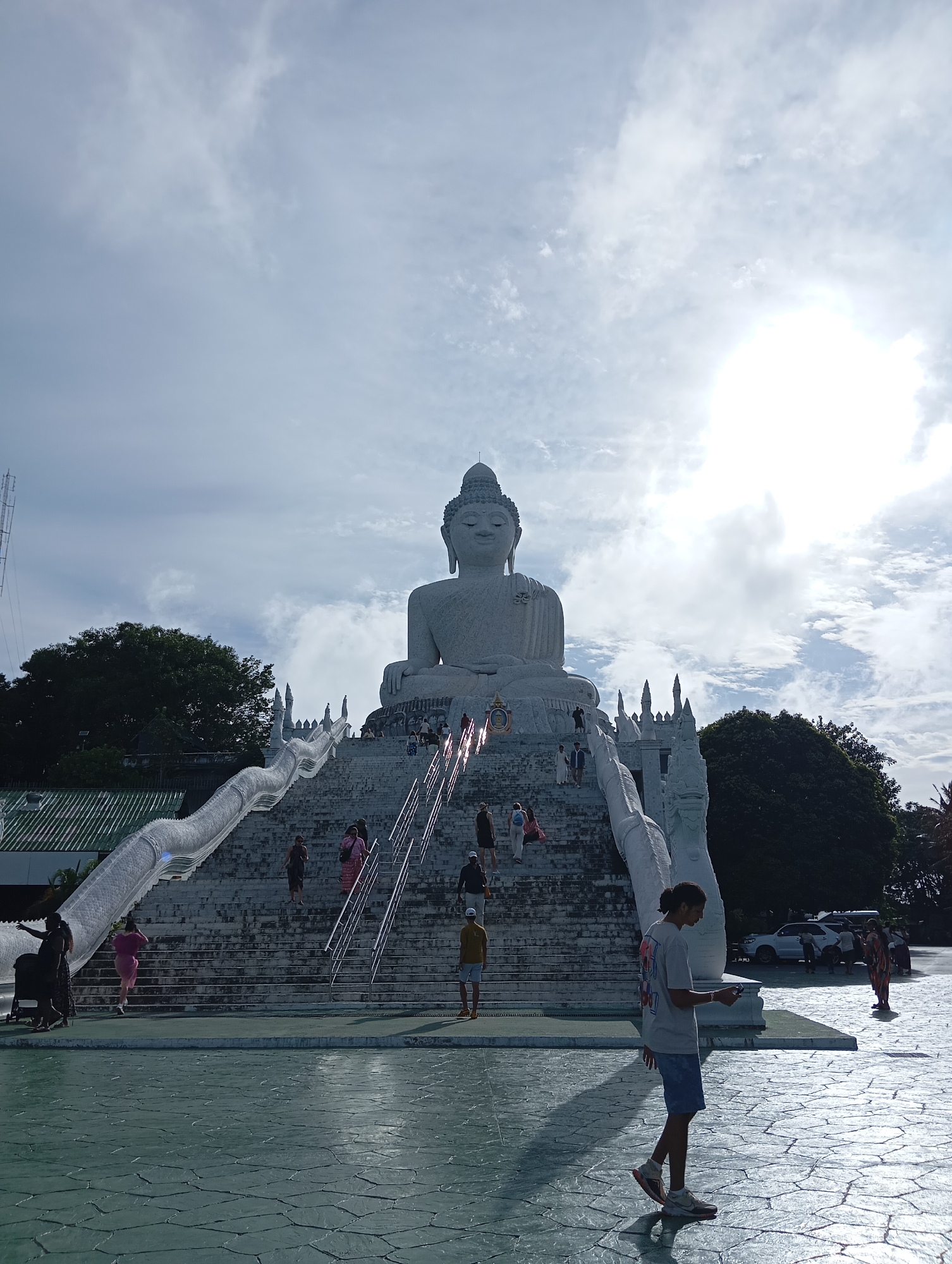 The image captures the Big Buddha statue in Karon, Thailand, on a bright, partly cloudy day. The massive white statue is seated majestically at the top of a grand staircase, which is flanked by ornate, dragon-like balustrades. Several individuals are seen ascending the stairs, some individually, others in small groups. In the foreground, a young man with dark hair and a grey t-shirt walks across a patterned paved area, casting a long shadow. In the background, trees and a white car are visible. The sun is shining brightly, creating a halo effect in the sky.