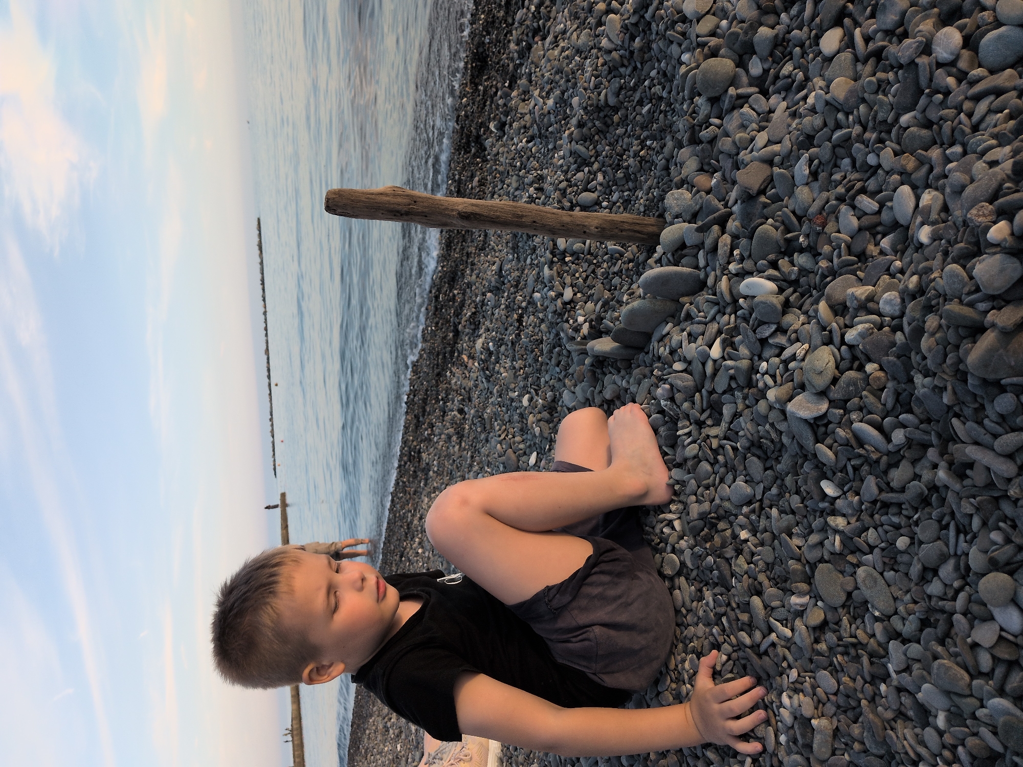 A young boy sits on a pebbly beach on the coast of Sochi, Russia, during the late afternoon or early evening. The boy, appearing to be between five and eight years old, has short, light brown hair and is dressed in a black short-sleeved t-shirt and dark grey shorts, with bare feet. He is positioned on the ground with his left arm extended to his side, hand resting on the dark grey and black pebbles. His legs are bent, and his gaze is directed downwards and to his right, indicating focus on something immediately in front of him on the ground or a contemplative state. To his right, a curved arrangement of several flat, upright pebbles suggests a structure he may have built. Adjacent to this stone arrangement, a piece of weathered, dark brown driftwood stands vertically in the pebbles. The calm, dark blue-green sea laps gently at the shore immediately behind the driftwood and the boy. In the distant background, several figures of people are visible near what appear to be rock breakwaters or piers extending into the sea from the left. The horizon is flat, and the sky above is a pale blue with scattered, wispy white clouds, illuminated by the soft light of the low sun.