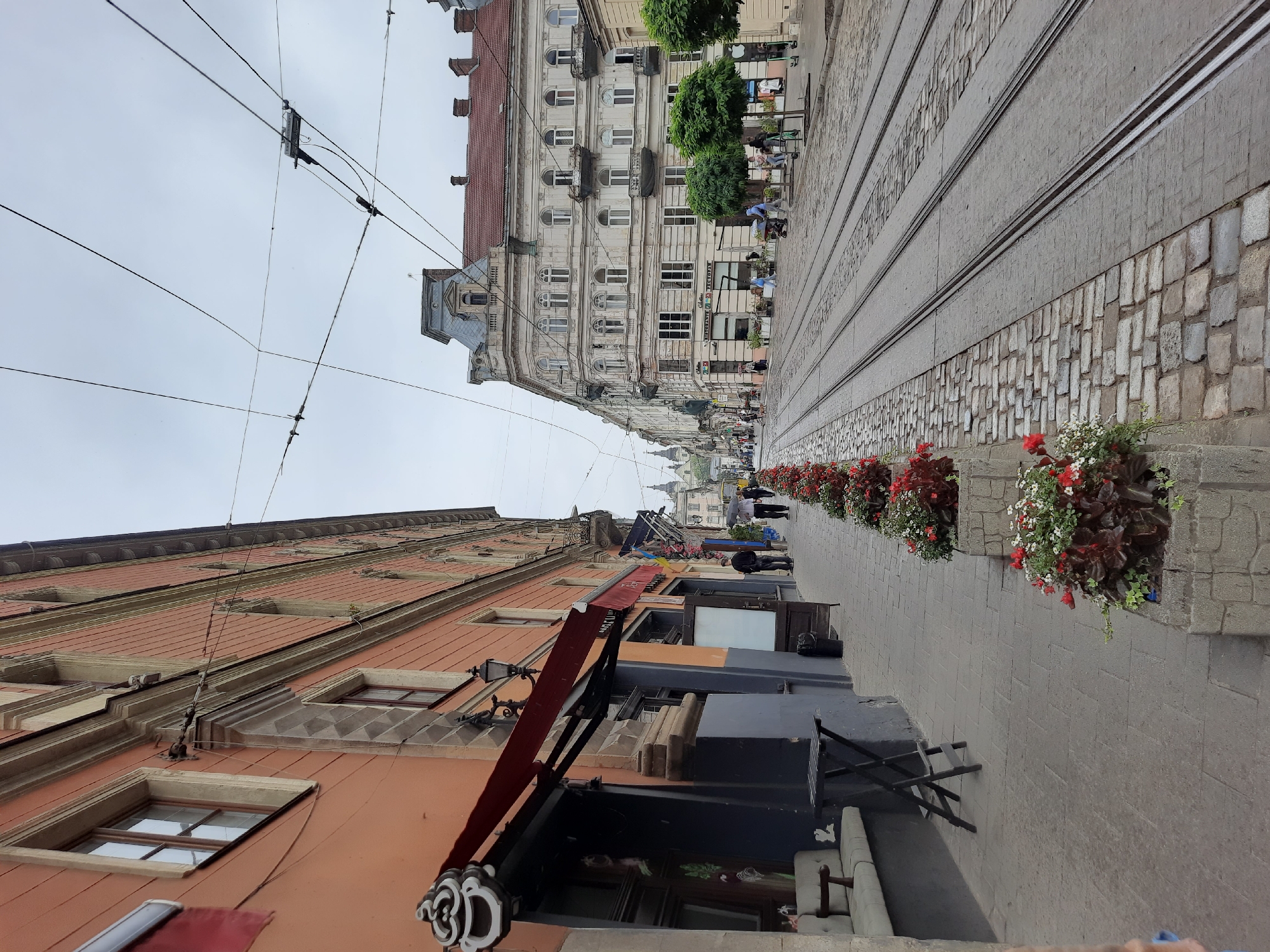 The image depicts a street scene in Lviv, Ukraine, featuring historic architecture and active tram lines. On the left, a building with an orange facade displays a red awning over a storefront with a decorative sign. A bench and a small table are placed on the cobblestone sidewalk in front of the establishment. Adjacent to this are several planters filled with red and white flowers.
The street itself is paved with cobblestones and bisected by multiple sets of tram tracks that extend into the distance. On the right side of the street, a grand, ornate building with multiple windows and balconies stands out. Several trees are planted along the sidewalk in front of this building.
Several individuals are visible in the scene. Two people walk on the sidewalk, one holding an umbrella, suggesting possible light rain or overcast conditions. Others are seen further down the street, some appearing to be patrons of businesses or pedestrians. The sky is overcast, contributing to a diffused lighting. The overall impression is of a lively, historic urban environment.