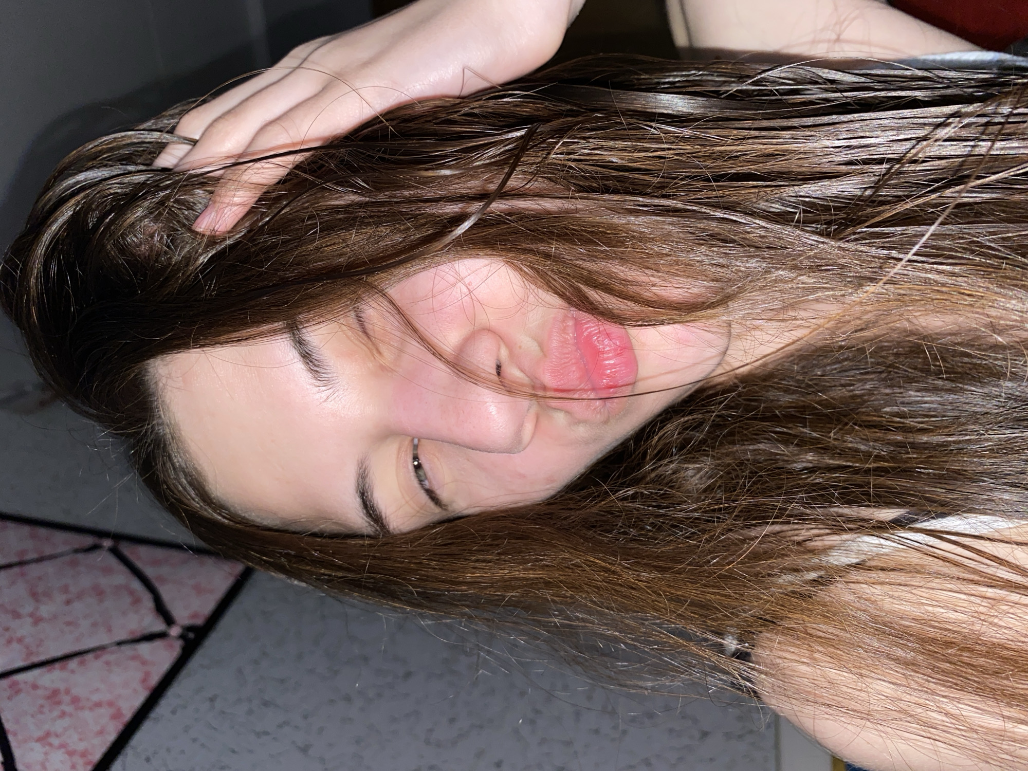 A young person with wet, dark brown hair is lying down with their head tilted. One hand is in their hair. Their eyes are closed and their lips are pursed. The background is slightly out of focus, showing a patterned floor and wall. There is no evidence to suggest this photo was taken in Votkinsk, Russia.