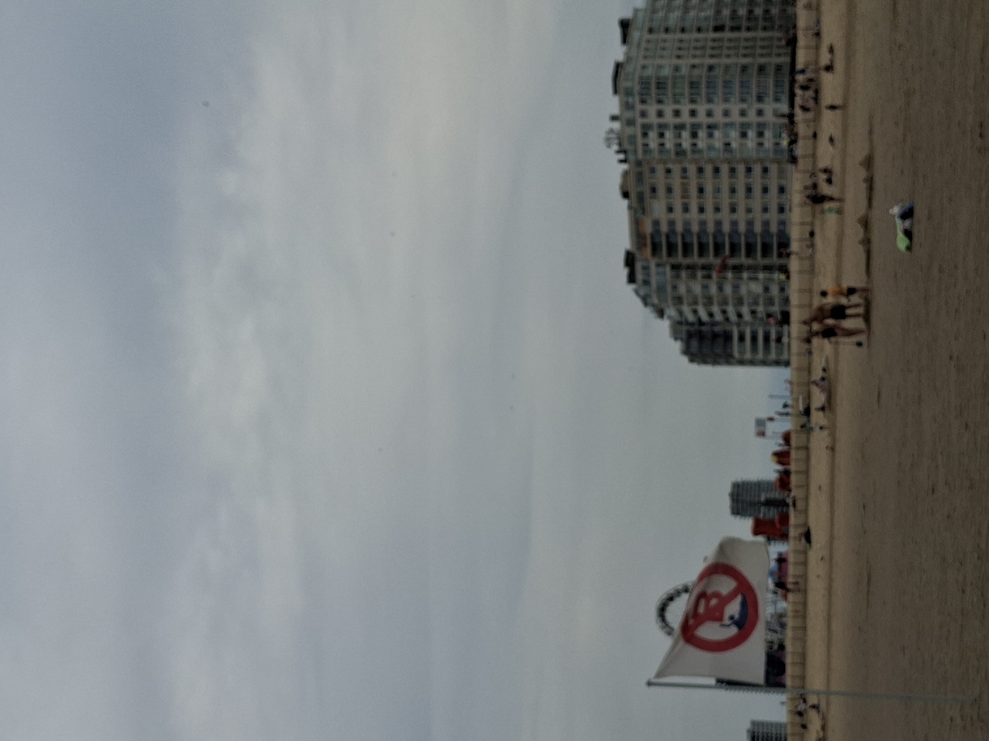 The image depicts a cloudy day at the beach in Ostend, Belgium. In the foreground, a sandy beach is visible with a few scattered individuals and beachgoers in the distance. A white flag with a red circle and a diagonal line across a silhouette of a surfer is prominently displayed on a flagpole. In the background, large apartment buildings line the coastline, characteristic of the Ostend promenade. A Ferris wheel can be seen to the left of the apartment buildings, suggesting the presence of an amusement area. The overall atmosphere is one of a typical seaside town on an overcast day.