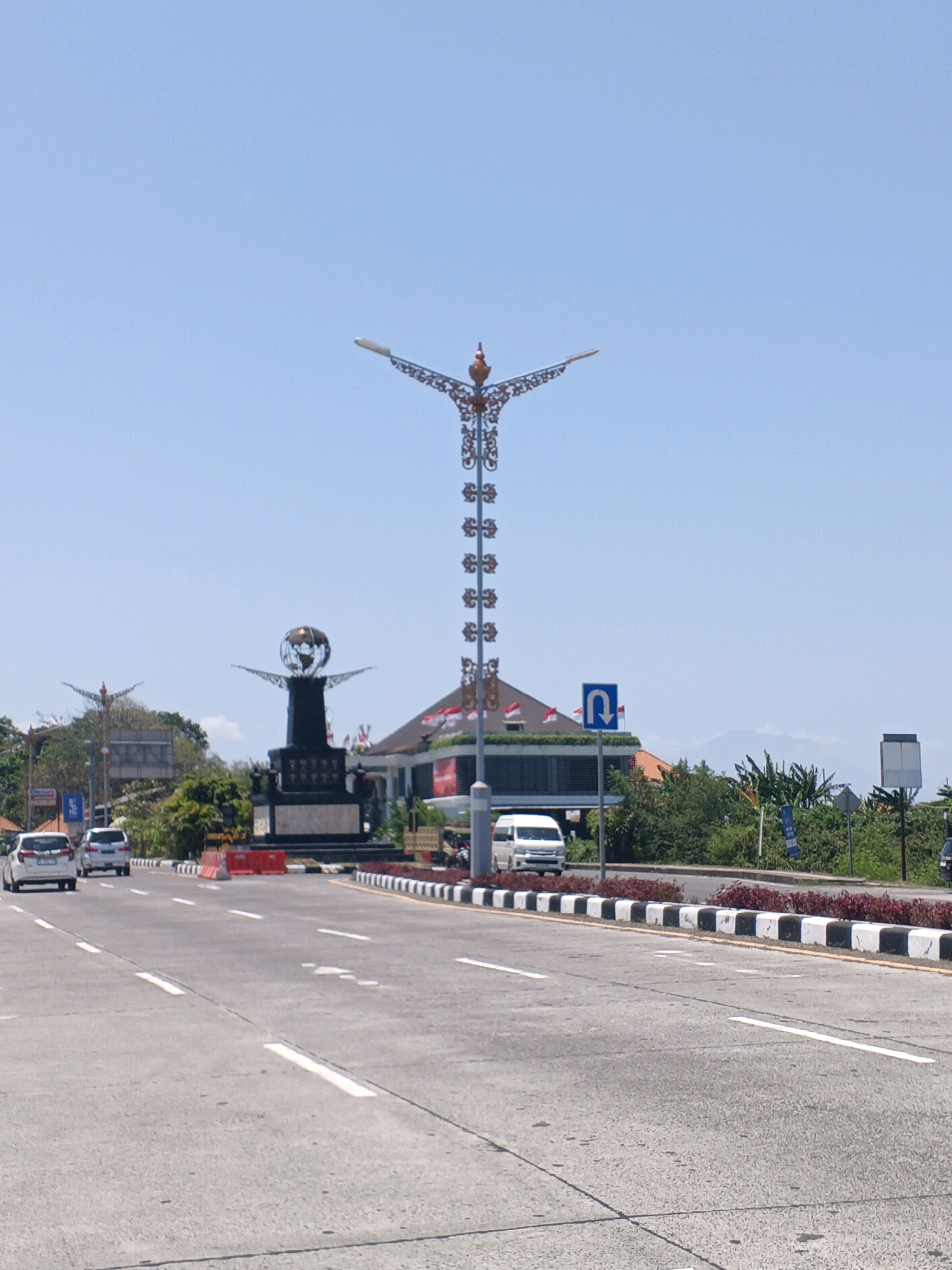 A bright, sunny day illuminates a wide asphalt road in Denpasar, Indonesia. The road features multiple lanes marked with white lines and a decorative black and white striped barrier lining the side. On the right side of the road, a white van drives away from the camera. Further in the distance, a large monument with a globe on top stands prominently. Ornate streetlights with intricate metalwork flank the monument. A building with a red and white flag on its roof is visible behind the monument. The sky is clear and blue. The scene appears to be a busy street with a notable landmark.
