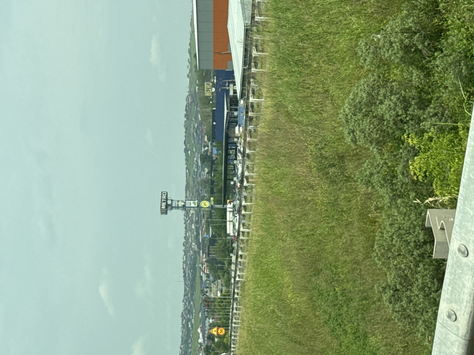 The image shows a wide shot of a commercial area on the outskirts of Solontsy, Russia. In the foreground, there is tall, green grass and some shrubbery. A concrete barrier with visible bolts runs along the bottom of the frame. In the middle ground, a highway or major road is visible with a white barrier separating it from the grassy area. Several cars and a truck are parked in a lot beyond the road, suggesting a commercial establishment. A large sign for "METRO" is prominently displayed on a tall pole, indicating a Metro Cash & Carry store. Adjacent to this, there is a large building with an orange and gray facade, and another building with a blue facade. In the background, a densely populated residential area with numerous houses and buildings stretches across rolling hills. The sky is overcast and a pale blue-green color. There are no people clearly visible in the image.