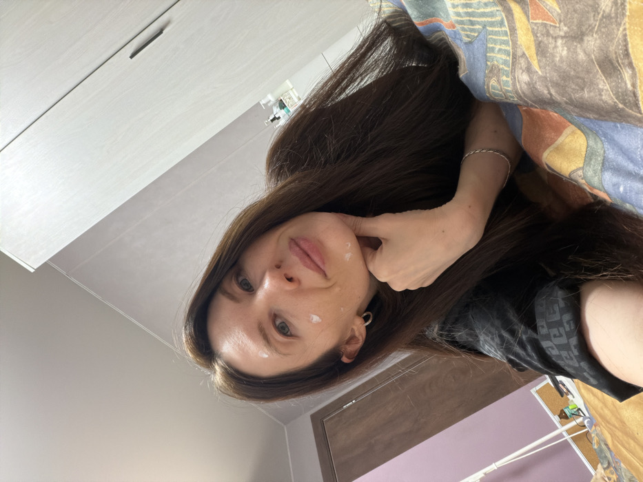 A woman with long, dark brown hair, wearing a black silk shirt, lies in bed. She is propped up with a colorful patterned pillow. She has cream on her forehead and cheek. She is touching her face. Her room has a white wardrobe on the left and a brown door on the right. A silver bracelet is visible on her wrist. The photo has a slightly tilted perspective.
