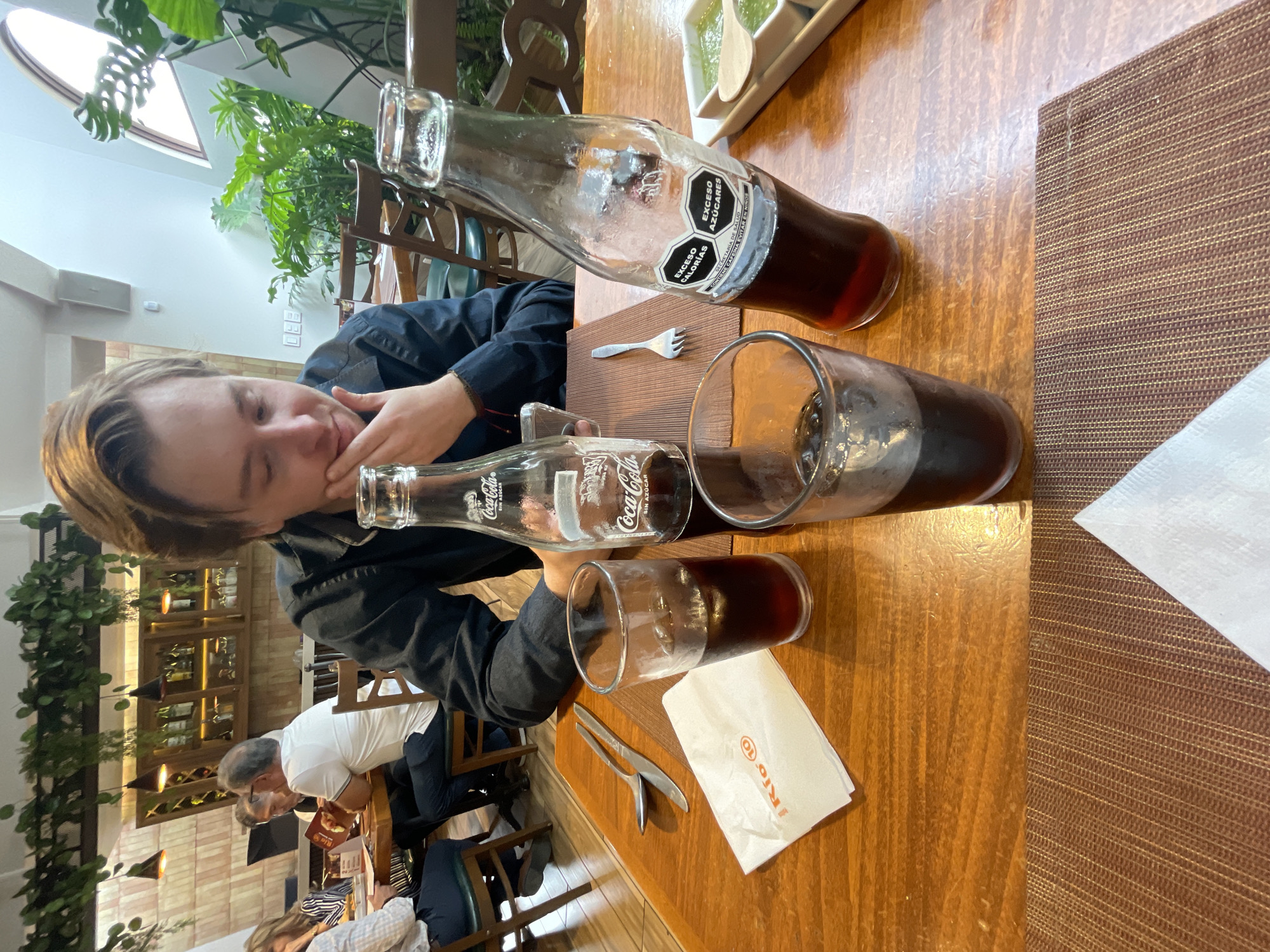 A young man with light brown hair is seated at a wooden table in what appears to be a restaurant. He is wearing a dark blue button-down shirt and is looking down at the table, with his right hand resting on his chin. In front of him are two glasses filled with a dark liquid and ice, and two glass bottles of Coca-Cola. One bottle is closer to the foreground and is partially visible, while the other is further back. A white napkin with an orange logo, cutlery (a spoon and a knife), and a fork are also on the table. In the background, other patrons are seated at tables, and a shelf unit with bottles is visible. Plants are arranged in the environment, and part of a slanted ceiling with a skylight is also present. The overall setting suggests an indoor dining establishment in Cuautitlán Izcalli, Mexico, based on the presence of the "Exceso Calorías" and "Exceso Azúcares" labels on the Coca-Cola bottle, which are common in Mexico.