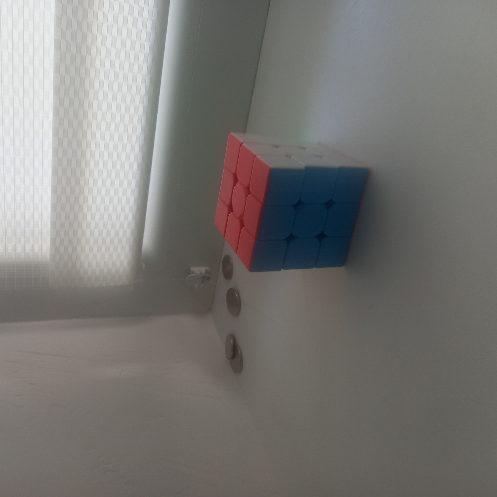 The image depicts a close-up, eye-level, slightly angled shot of a Rubik's Cube positioned on a flat white surface. The cube exhibits red and blue colored faces, with some white faces also visible. To the left of the cube, several silver coins are scattered on the same white surface. In the background, a window with horizontal blinds is visible, suggesting an indoor setting. The lighting appears to be natural, coming from the window, casting soft shadows. The overall environment suggests a quiet, possibly domestic setting. The scene is static and does not depict any activity or interaction. No people are present in the frame. The location in Cepeleuți, Moldova, is inferred from external context, but is not visually evident in the image itself.