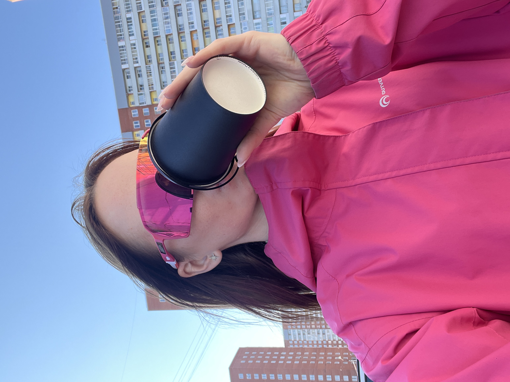 A young woman is captured at an outdoor location in Berezniki, Russia. The photo is taken from a lower angle, looking up at her. She is holding a black cylindrical container, possibly a cup, close to her face. She has on pink tinted sunglasses and wears a bright pink jacket. Her hair is dark and flows past her ear, where a star-shaped earring is visible. In the background are tall apartment buildings, typical of urban Russian architecture. The sky above is a clear, bright blue. The overall impression is that she is enjoying a drink outdoors in a city setting.