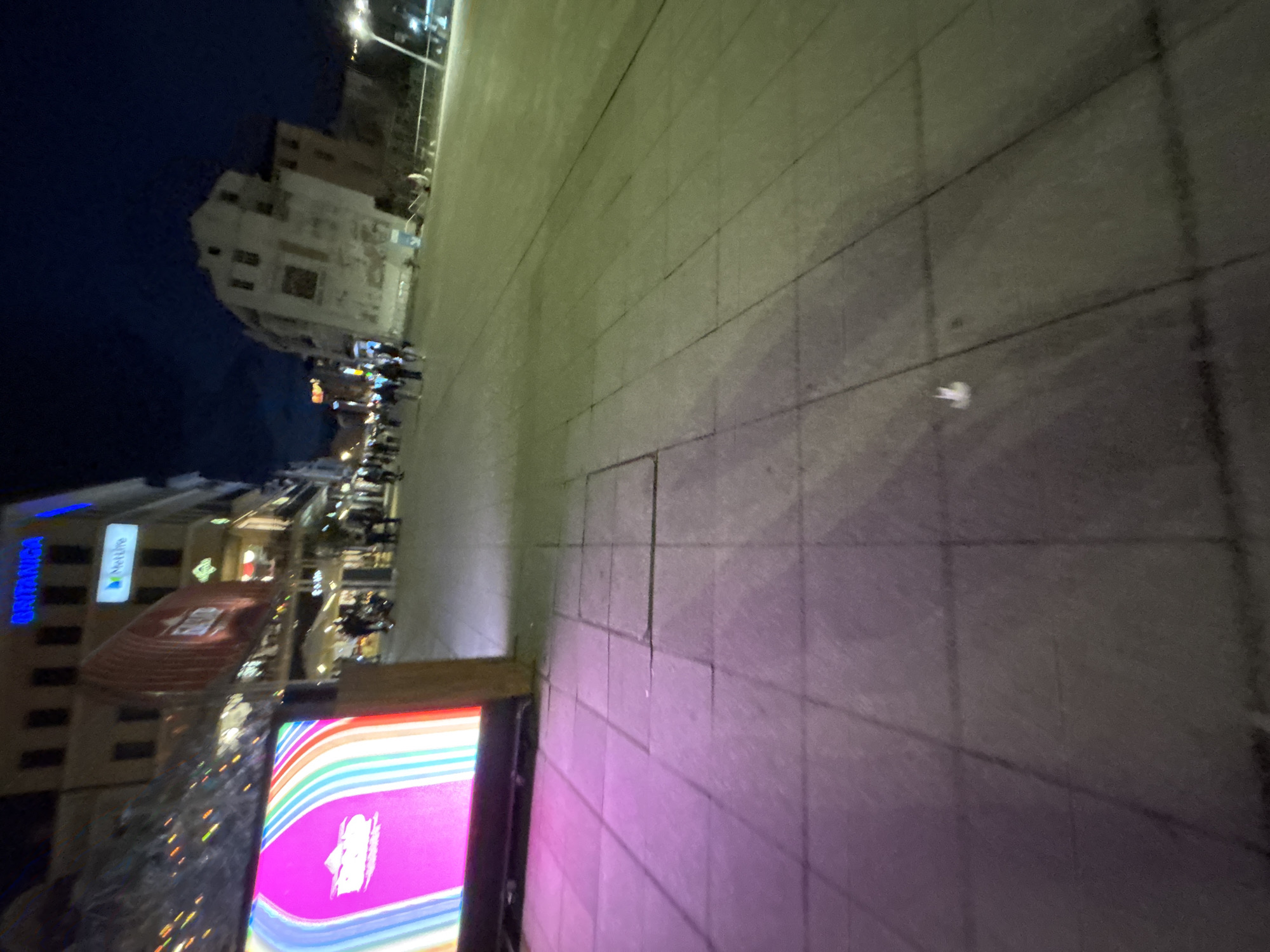 The image depicts a street scene at night in Plovdiv, Bulgaria. A multi-story building with a "Britanica" sign and "MetLife" logo is visible on the left. A building with a red awning and tiered seating area is next to the building. To the right is an open plaza with stone paving. In the distance, a historic building is visible against the night sky. People are walking on the street in the background. A colorful advertising display stands in the foreground.