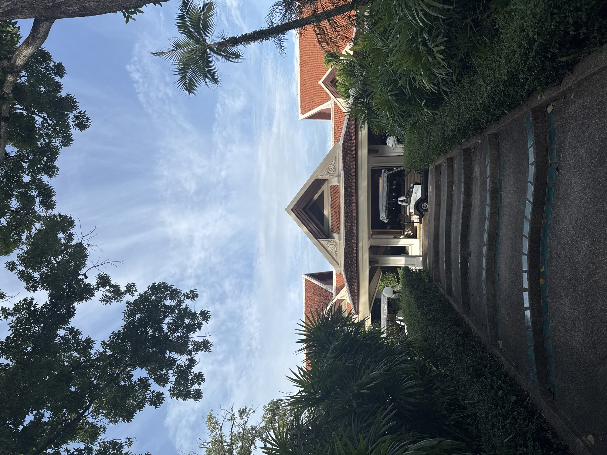 A wide shot captures the exterior of a building, likely a resort or hotel, in Ko Samui District, Thailand. The building features a distinctive traditional Thai architectural style with prominent red-tiled roofs and ornate gable details. The entrance is approached by a set of wide, stone steps, bordered by lush green foliage and manicured hedges on either side. A golf cart is parked near the entrance. Overhead, large tree branches frame the scene, with a clear blue sky and wispy white clouds visible in the background. A single palm tree stands tall to the right of the building. The lighting suggests it is daytime, possibly late afternoon, given the shadows cast. No people are clearly visible, but there may be individuals inside the building or out of frame.
