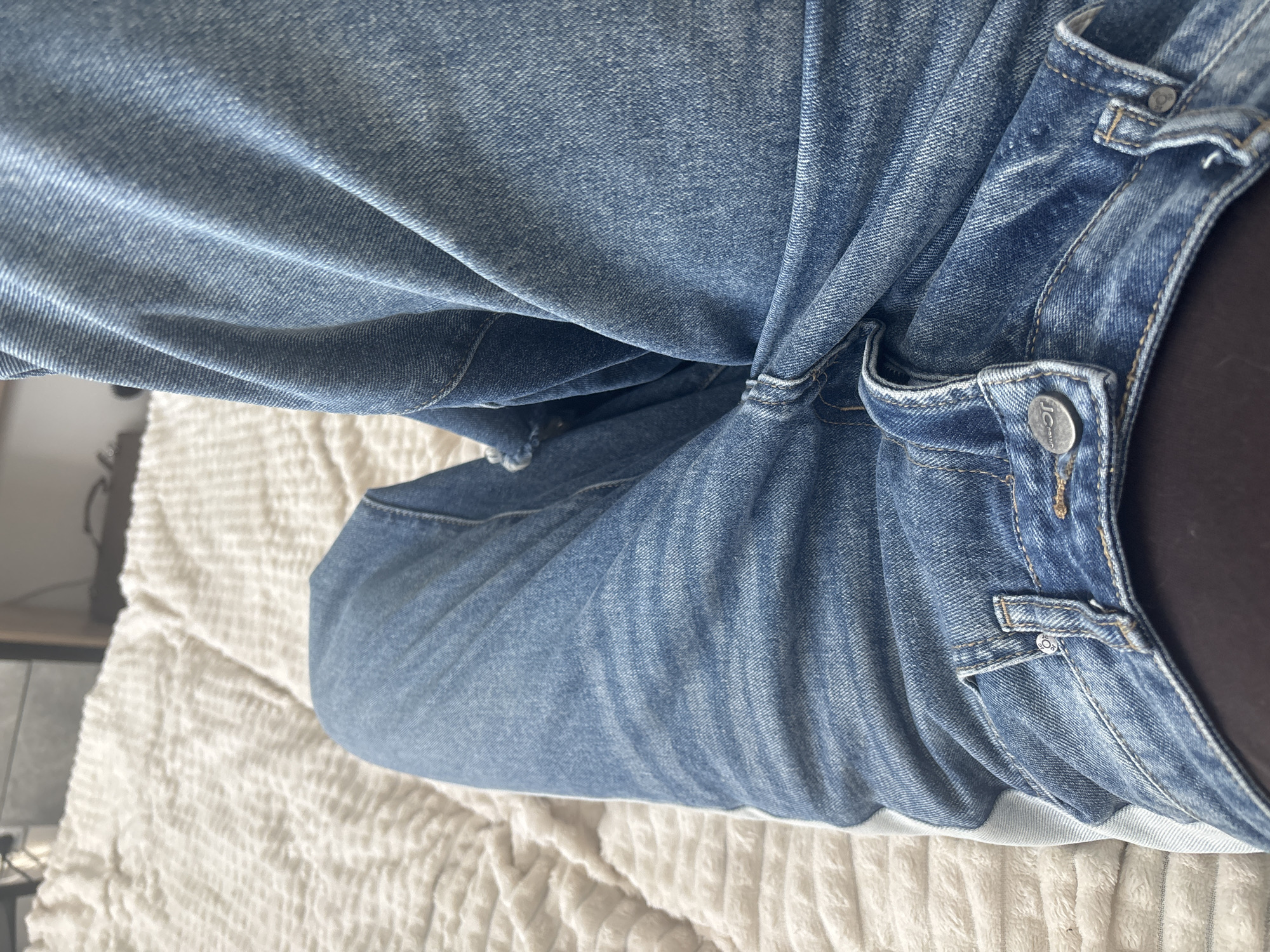 The image shows a close-up view of a person wearing blue jeans, lying on a white, textured bedspread. The focus is on the denim fabric, the waistband, and the button closure of the jeans. The button appears to have a logo, possibly "J. Crew". The person's skin is visible near the waistband. The environment is limited to the bed and a small portion of a bedside table in the background.
There is no information in the image that suggests the location is Irinovka, Russia.