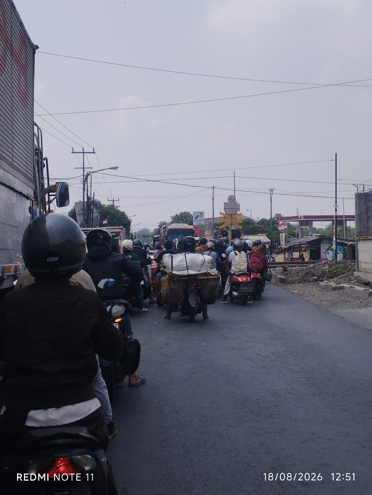 The image depicts a traffic jam on a city street, likely in Karawang, Indonesia, given the presence of "REMI NOTE 11" and "18/08/2026 12:51" timestamp at the bottom. The scene is dominated by a multitude of motorcycles carrying one or two passengers each, creating a dense flow of traffic. Many riders are wearing helmets, and some motorcycles are equipped with luggage racks carrying parcels. In the background, there's a large truck on the left, and further ahead, a gas station with a Pertamina logo is visible, alongside a small shack and a fence. The sky is overcast, and the road surface appears to be asphalt. The overall impression is one of congestion and daily urban life.